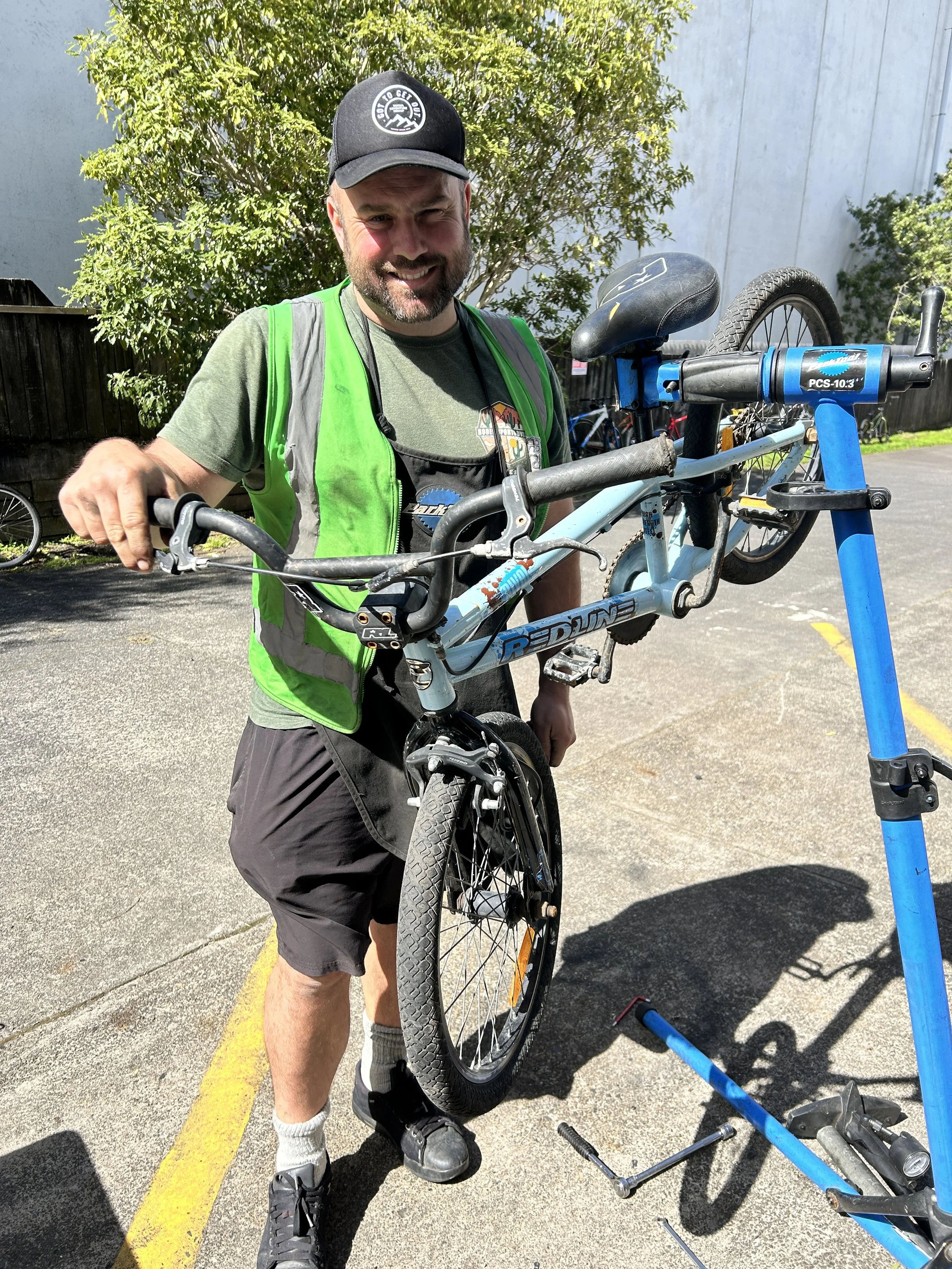 A man with a beard wearing a black cap, green safety vest, and gray t-shirt holds a bike upside down on a bike repair stand in an outdoor parking lot.