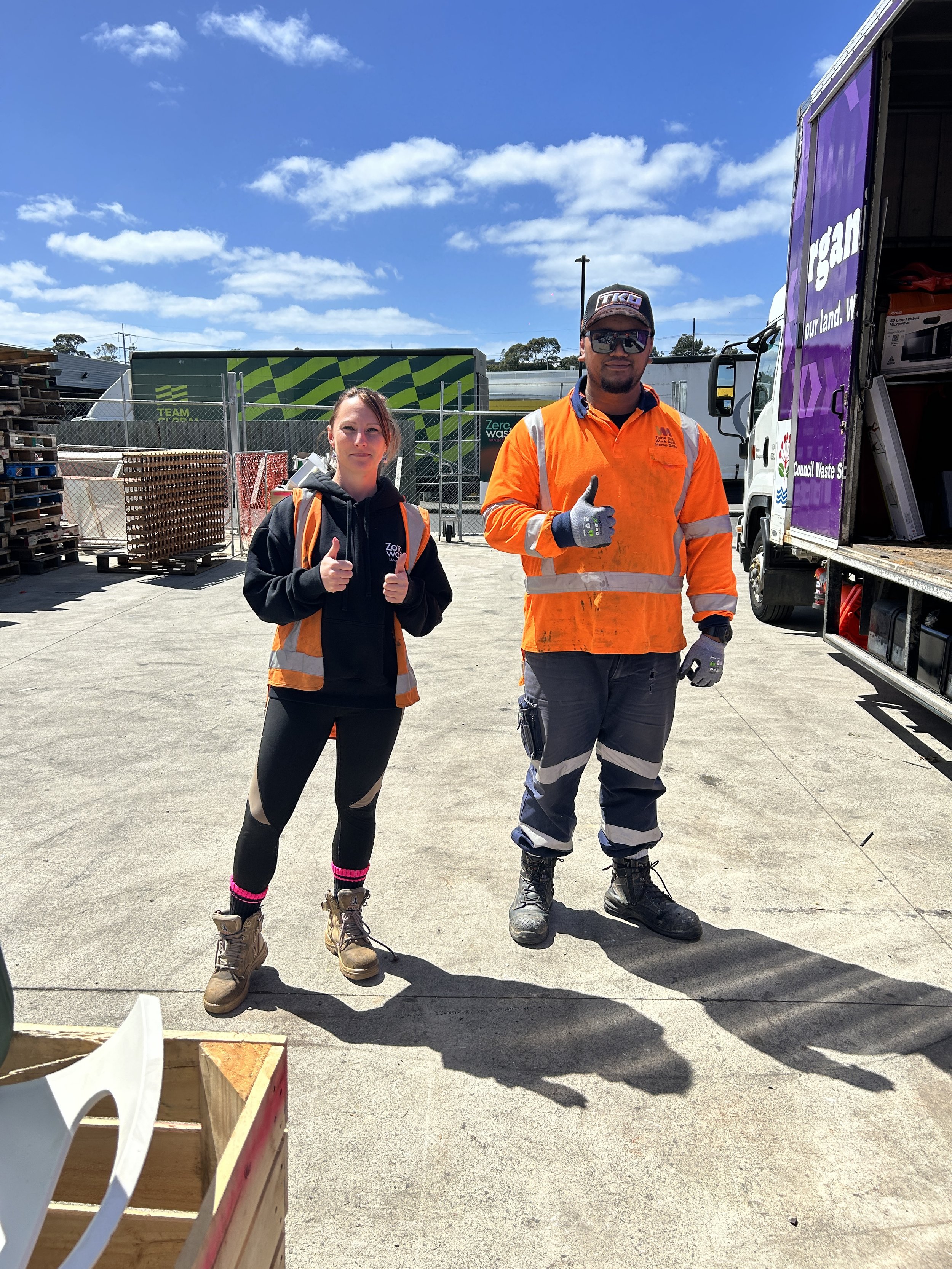 Two waste management workers, a woman and a man, wearing high-visibility orange safety jackets and boots, standing on a concrete surface next to a waste collection truck with purple and white branding, giving thumbs up. The background shows a chain-link fence, pallets, and a partly cloudy blue sky.