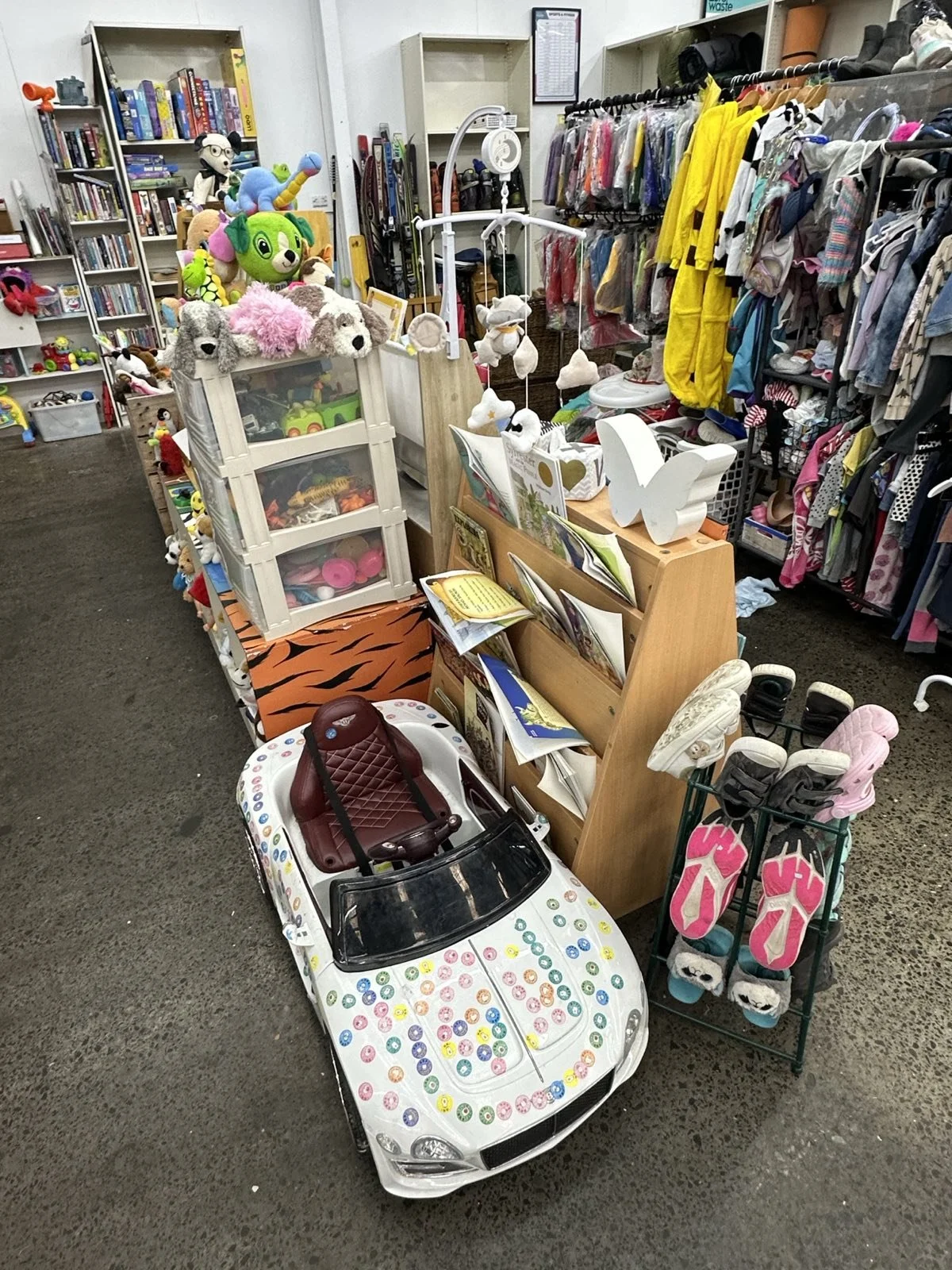 A toy store display featuring a miniature white race car with colorful stickers, a red seat, and a small black windshield, surrounded by plush toys, books, and children's clothing and footwear.