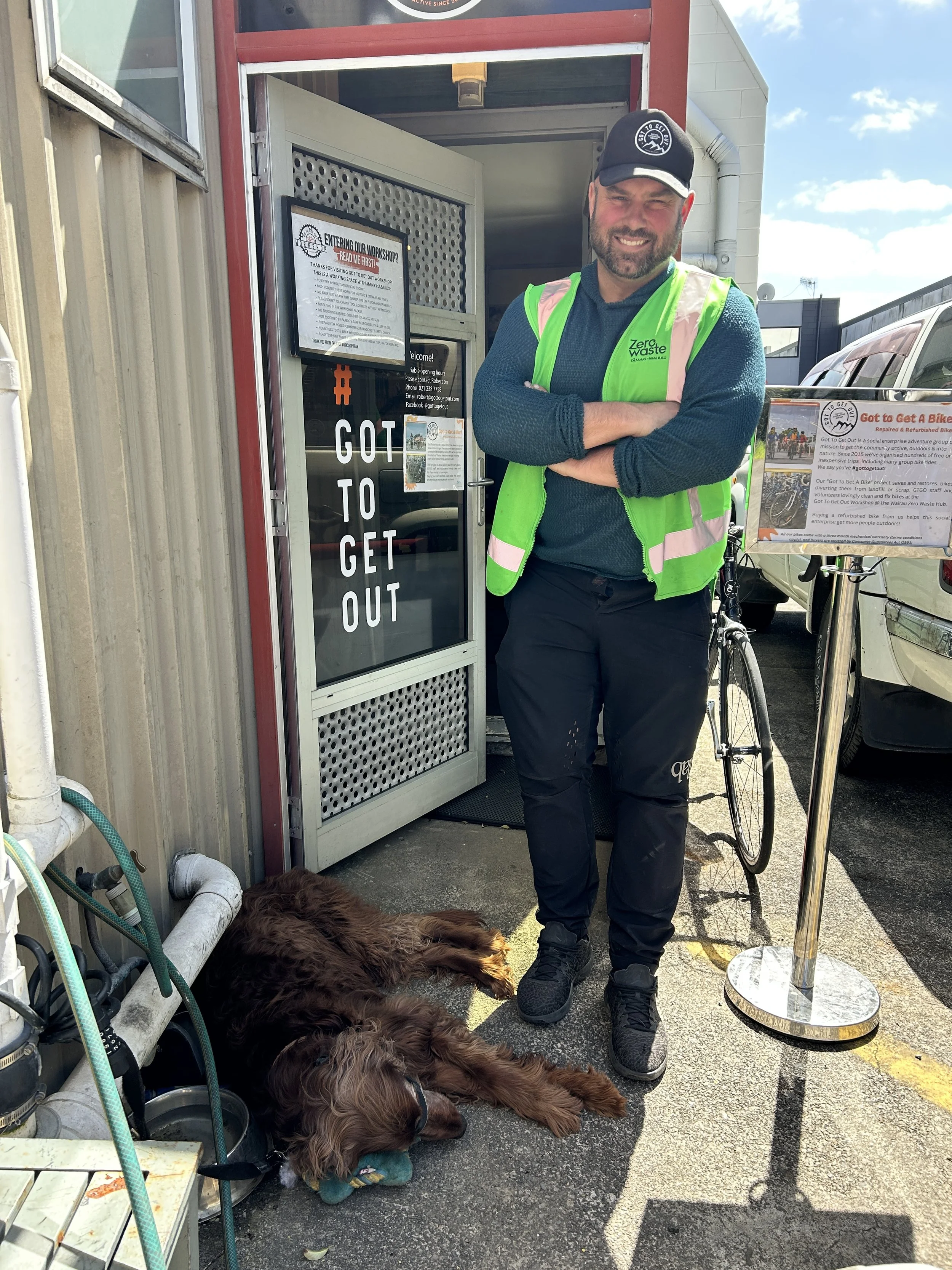 A smiling man wearing a neon green safety vest, black pants, and a black baseball cap standing with crossed arms next to a dog lying on the ground. The dog is a brown, long-haired breed resting with a toy in its mouth. They are outside a building with a sign that reads 'GOT TO GET OUT' and a poster about bike repair volunteer program.