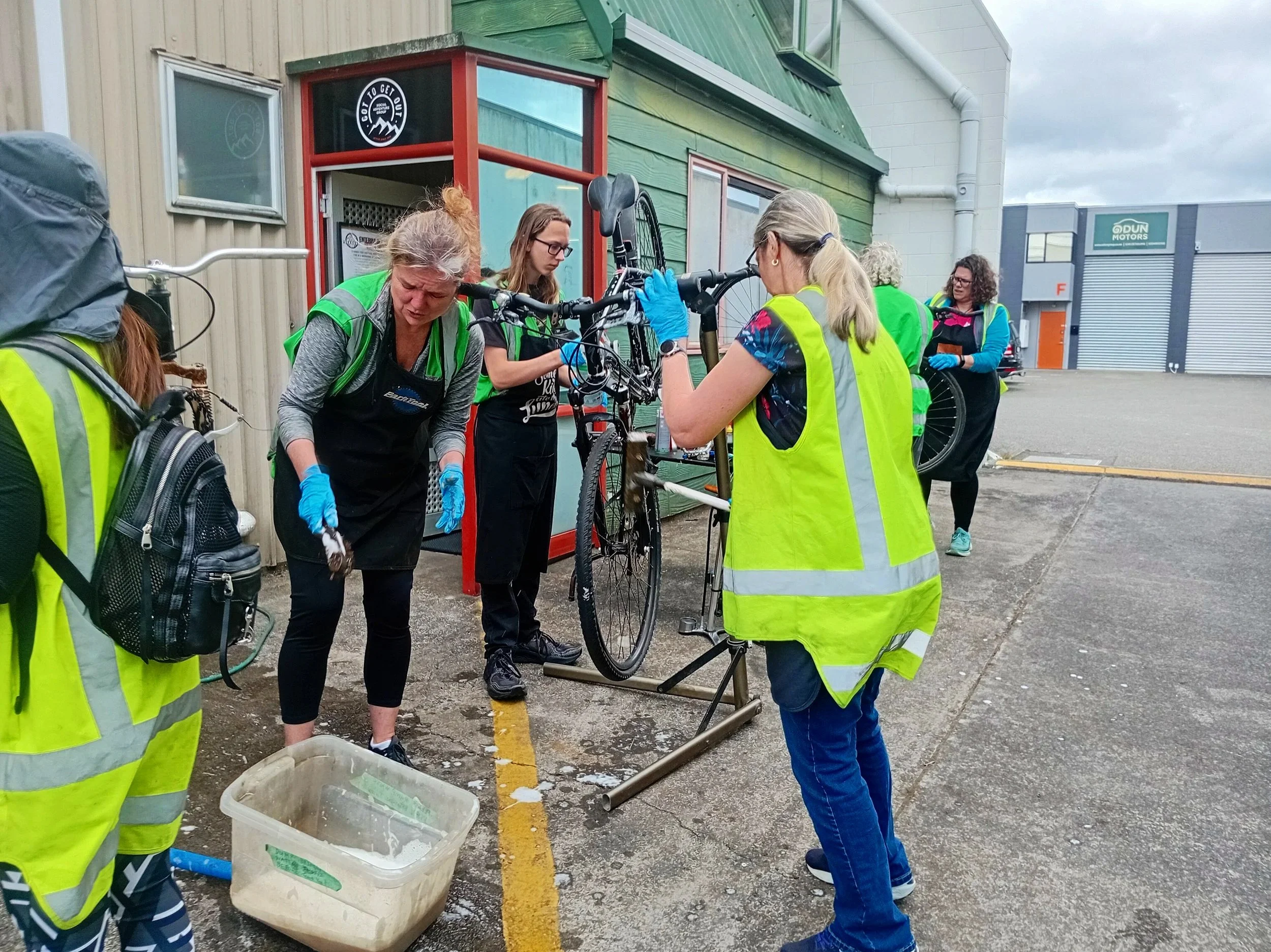 Group of people cleaning and repairing bicycles outside a building, some wearing high-visibility vests and gloves.