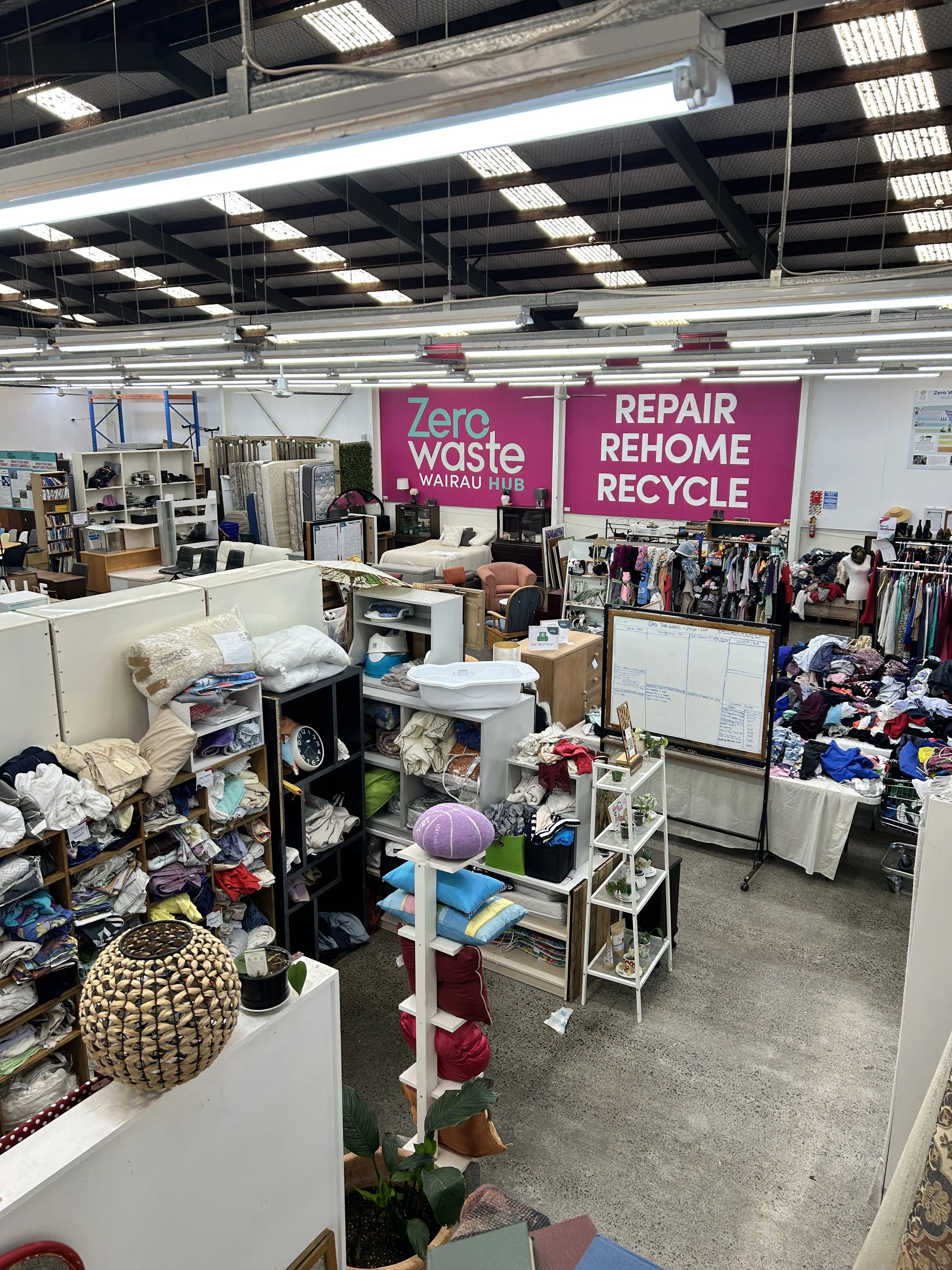 Interior of a thrift store with various household items, clothing, and furniture on display, and a large pink sign in the background that reads 'Zero Waste Wairau Hub' and lists services like repair, rehome, and recycle.