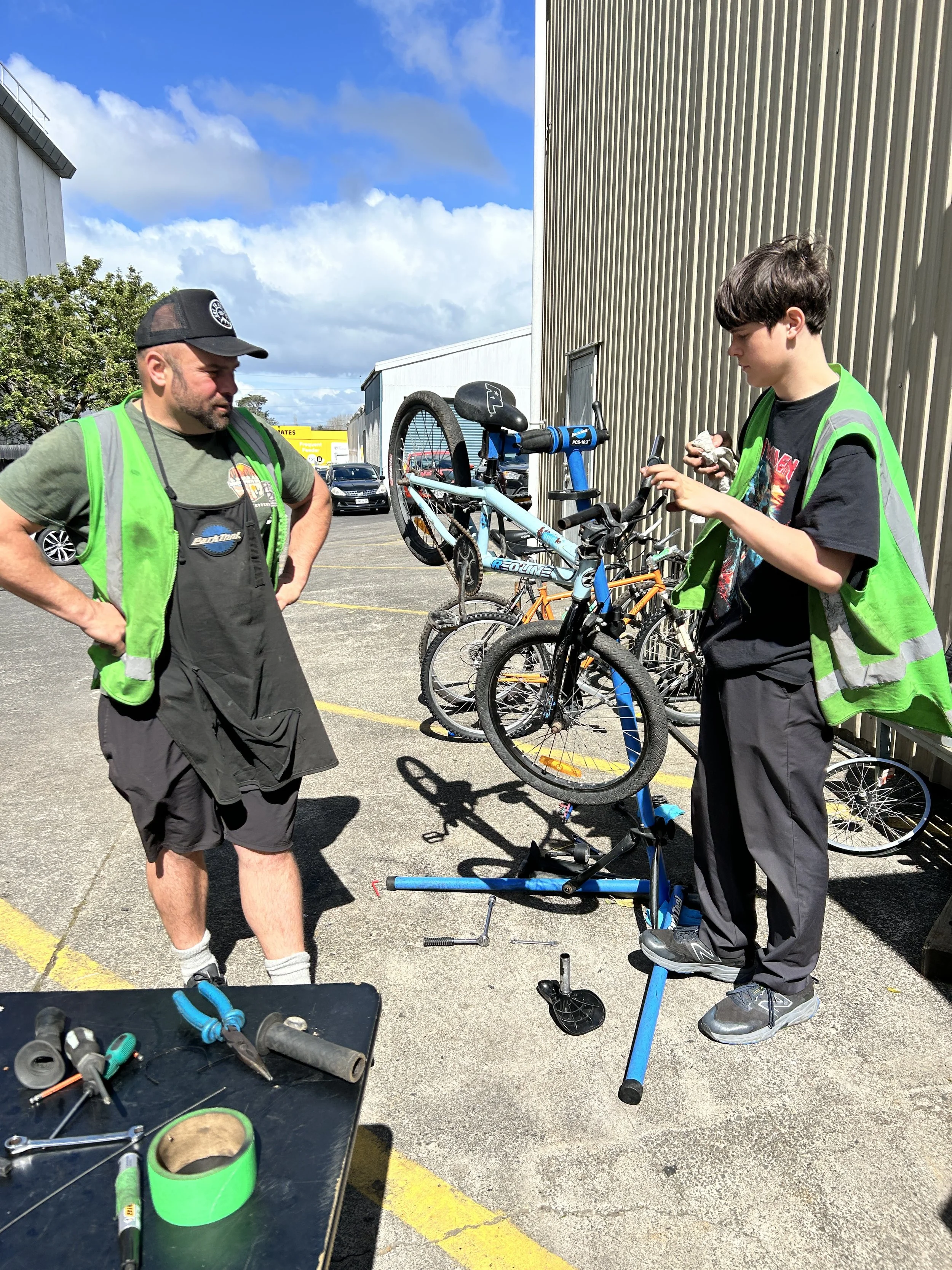 A young boy repairs a bicycle on a bike repair stand while an adult supervises. There are multiple bicycles hanging on a rack behind them. The scene takes place outdoors in a parking lot near a building under a partly cloudy sky.