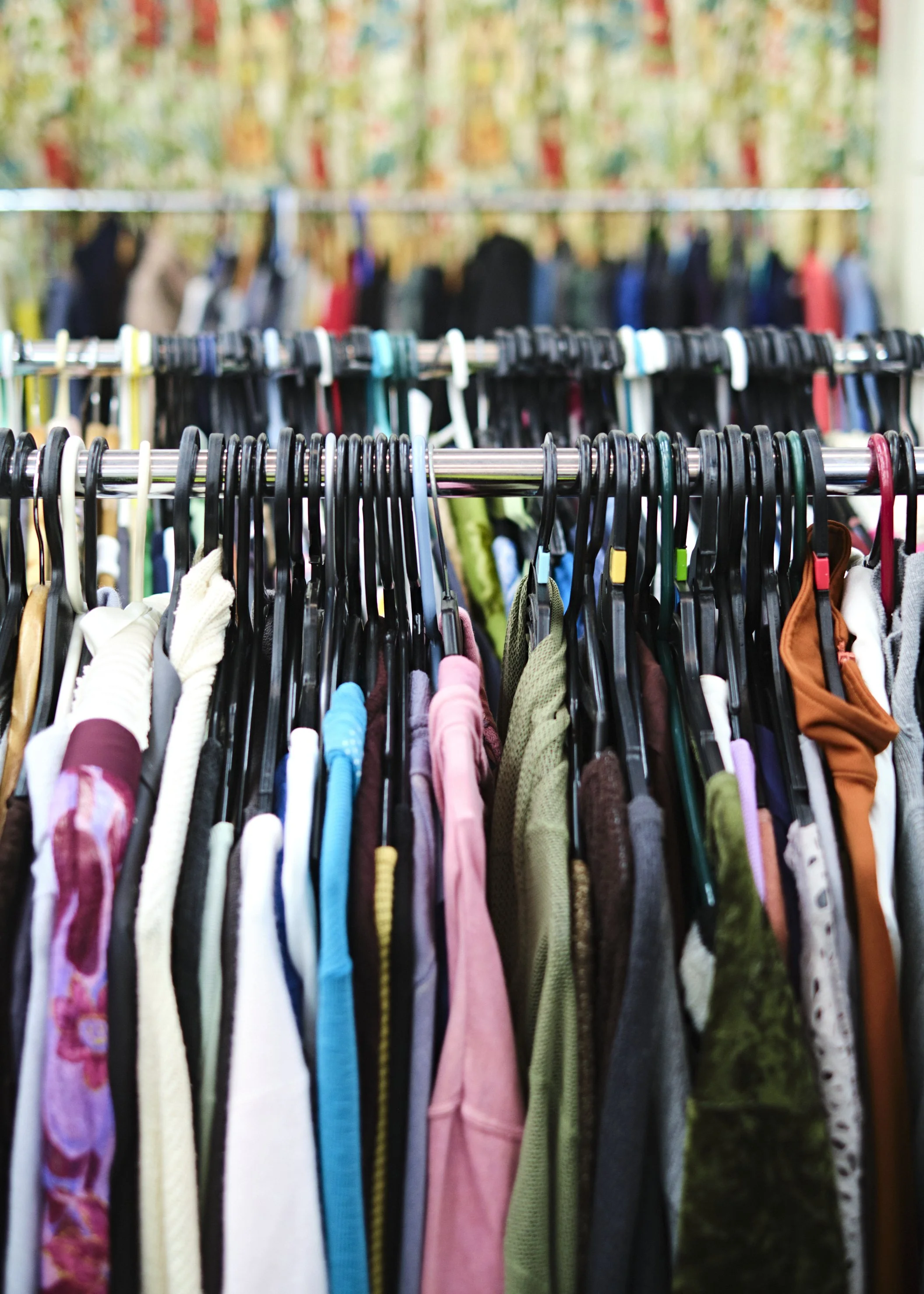 Clothes hanging on racks in a thrift or second-hand store, with various colorful garments on black plastic hangers.