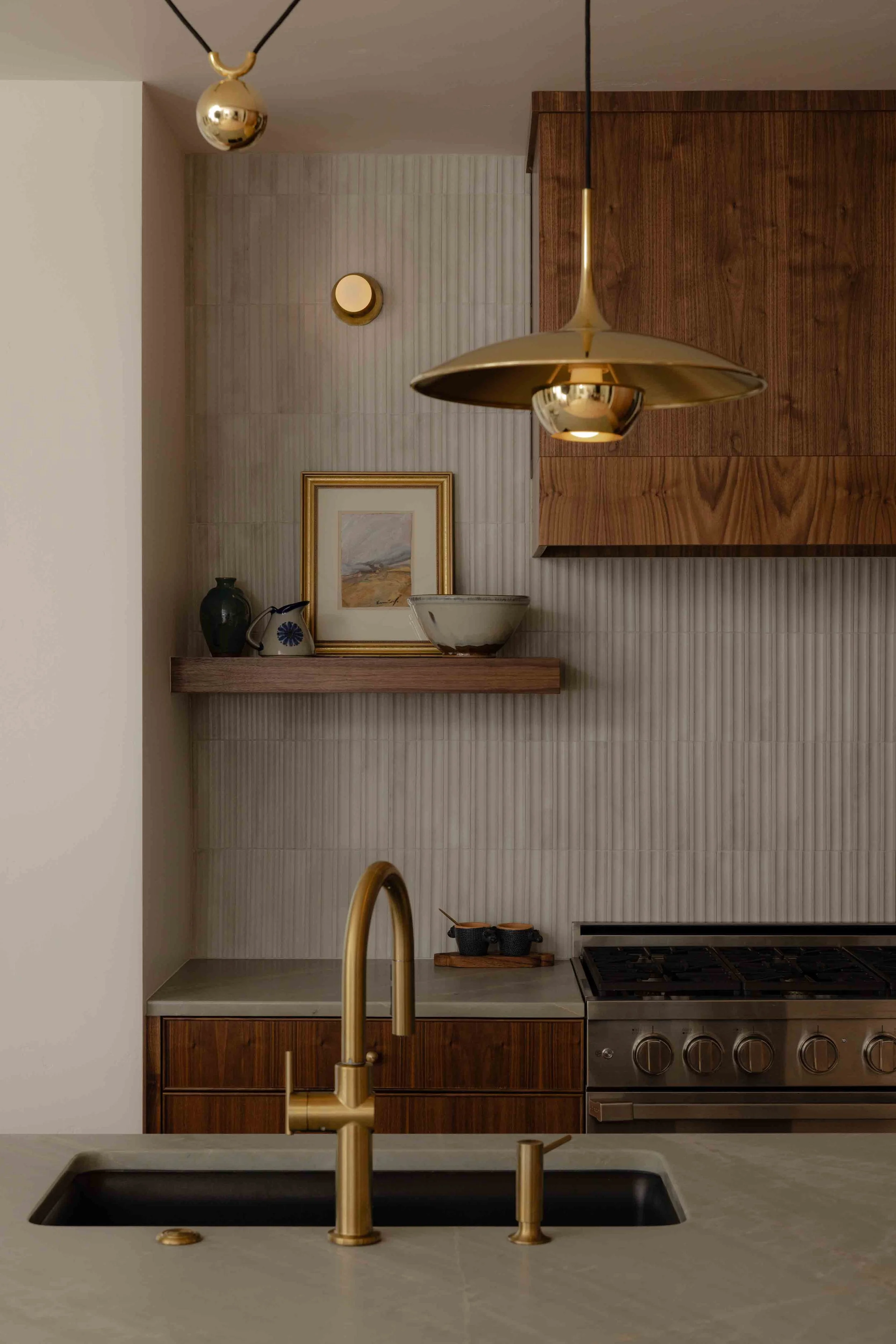 A modern kitchen with a gold sink faucet, a marble countertop, and wooden cabinetry. There are decorative items including a framed picture, vases, bowls, and hanging pendant lights with gold finishes.