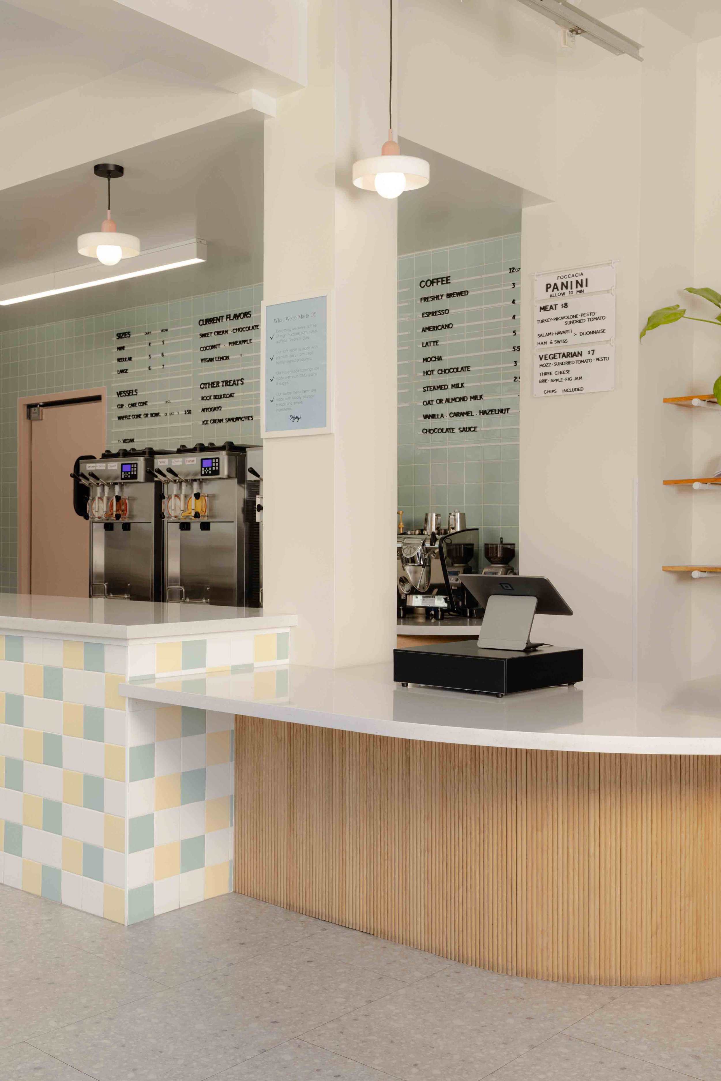 Interior of a modern coffee shop with a white counter, coffee machines, menu boards, and pendant lighting.