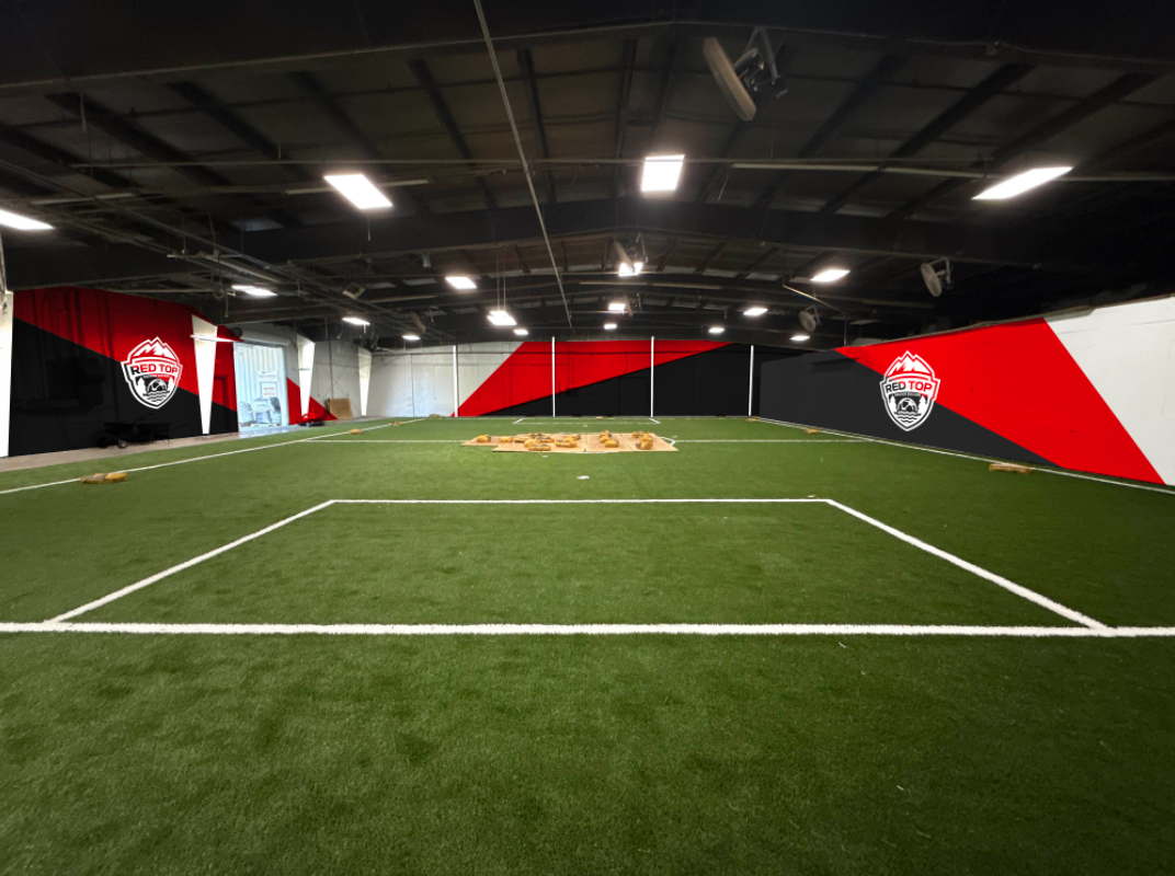 Indoor sports training facility with artificial turf, soccer goal, and wall murals with a red, black, and white logo reading "Red Top."