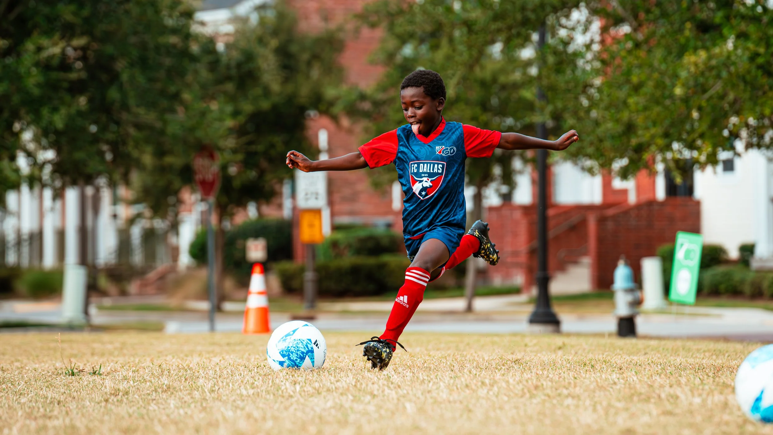 Young boy playing soccer outdoors on a grassy field, kicking a blue and white soccer ball while wearing a blue and red soccer uniform with a logo that reads 'FC Dallas', red shorts, and red socks.