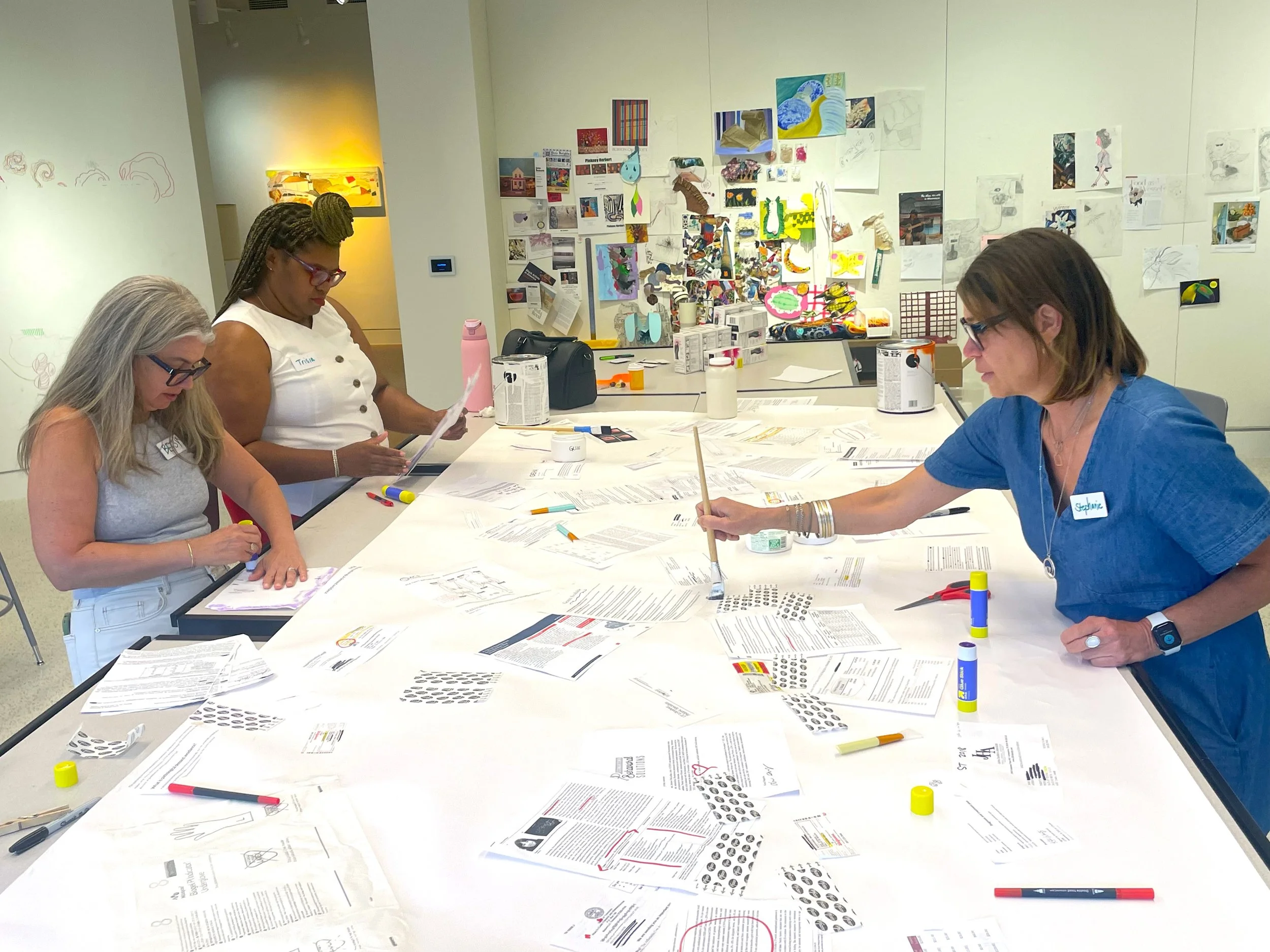 Three Gather participants work around a long table covered with medical paperwork and art supplies.