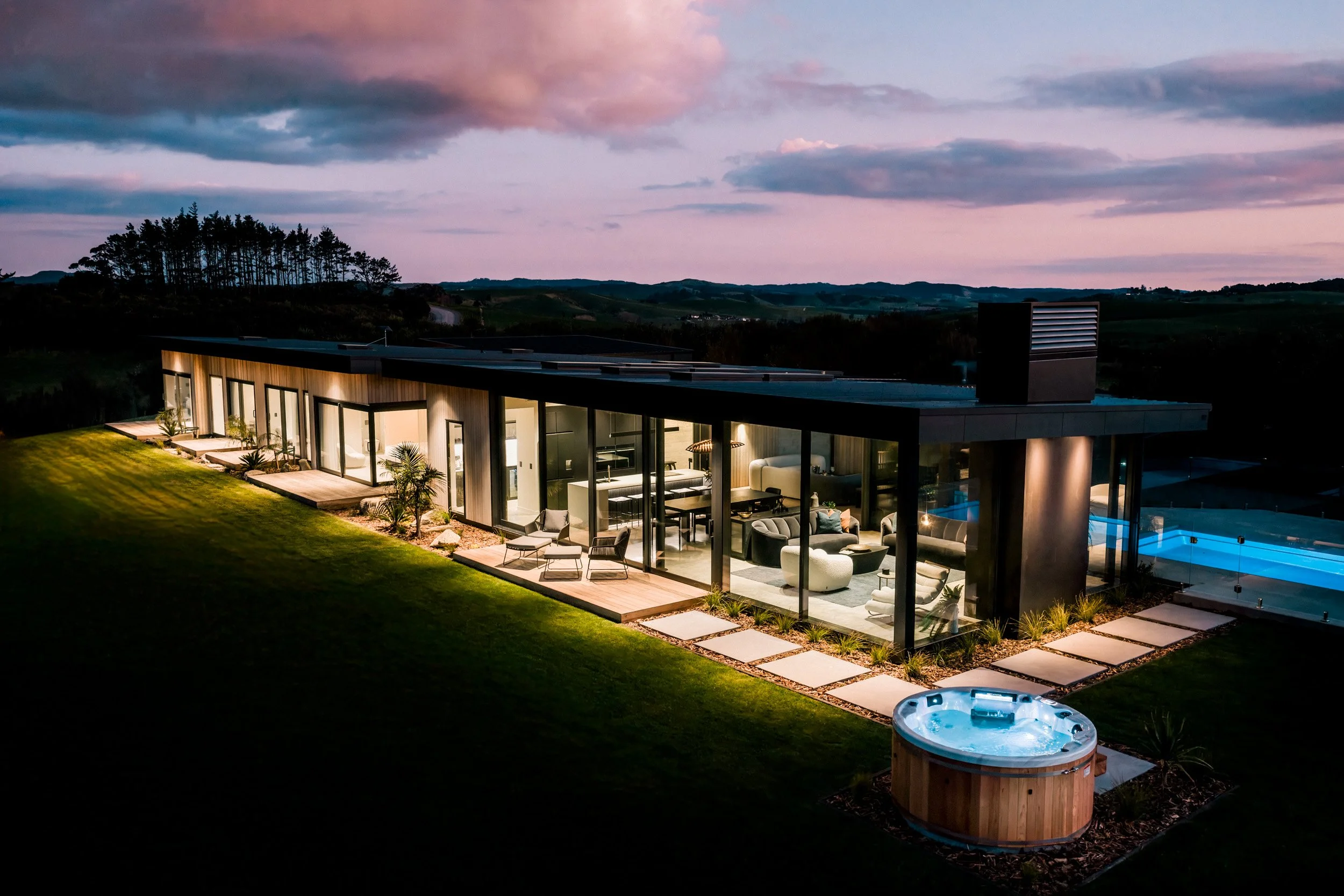 Twilight three-quarter angle exterior of Ocean View Te Arai showcasing seaward-facing decks, floor-to-ceiling glass revealing designer furniture and feature lighting, with an illuminated swimming pool and spa.