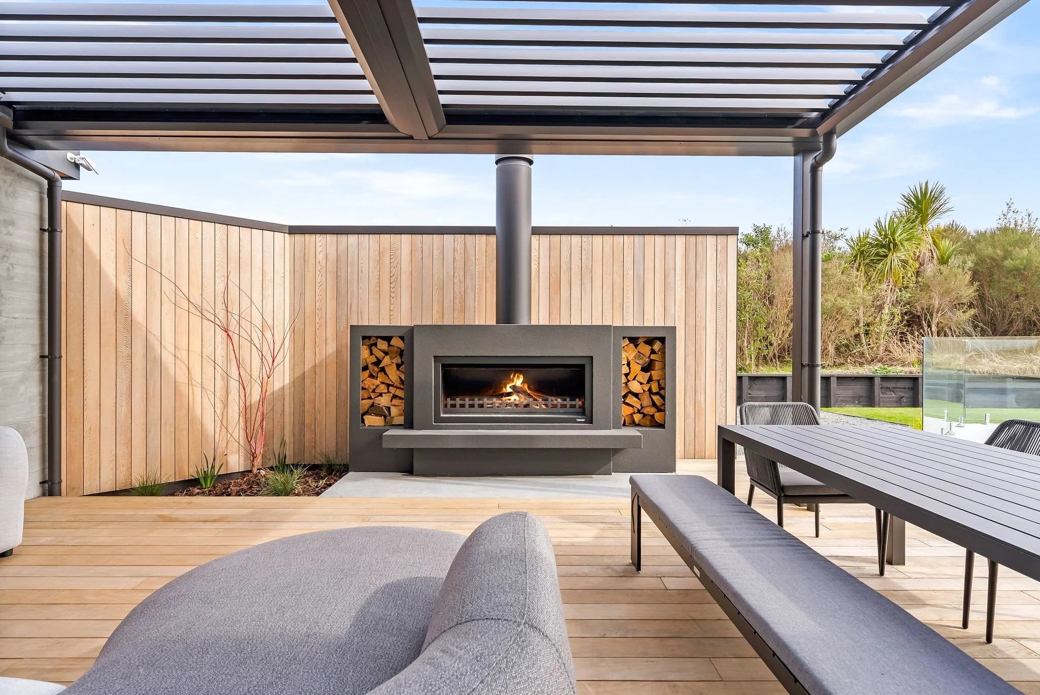Modern freestanding fireplace at the centre of a louvre-covered outdoor entertaining area at Ocean View Te Arai, featuring a contemporary lounge suite, dining table, and cedar feature wall.