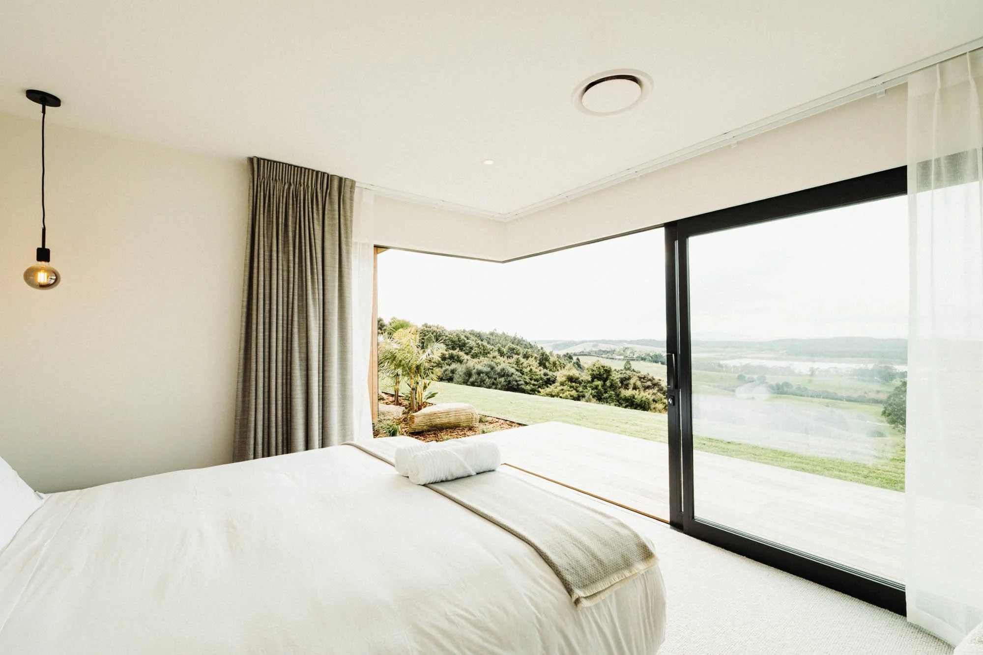 One of the two master bedroom suites at Ocean View Te Arai featuring corner sliding doors opening to expansive views of the Pacific Ocean, Hen & Chicken Islands, lakes, pine forest, and farmland, with a super king bed, premium bedding, and luxury fin