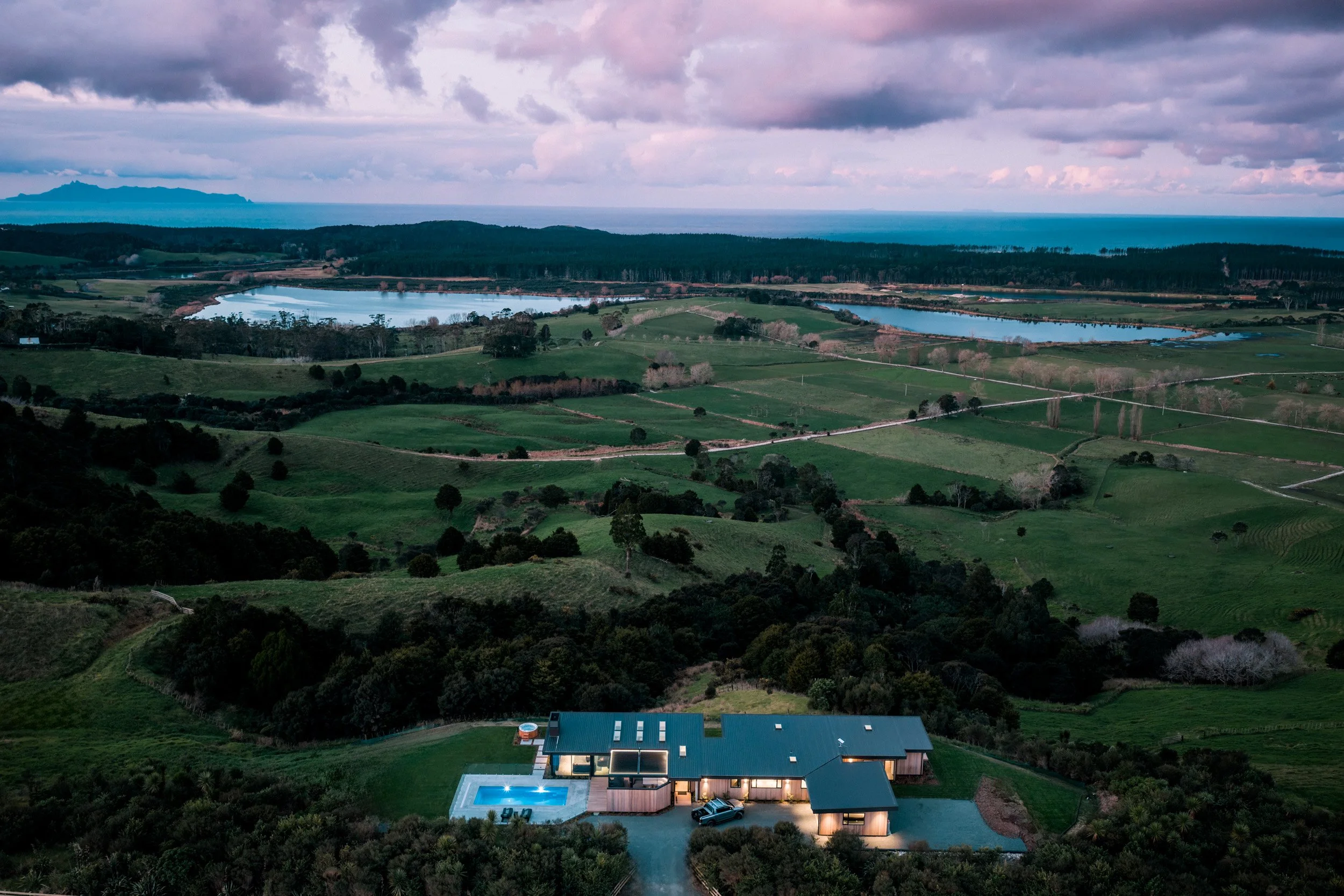 Twilight exterior of Ocean View Te Arai with illuminated swimming pool in the foreground, overlooking rolling farmland, lakes, Te Arai Links pine forest, the Pacific Ocean, and Hen & Chicken Islands.