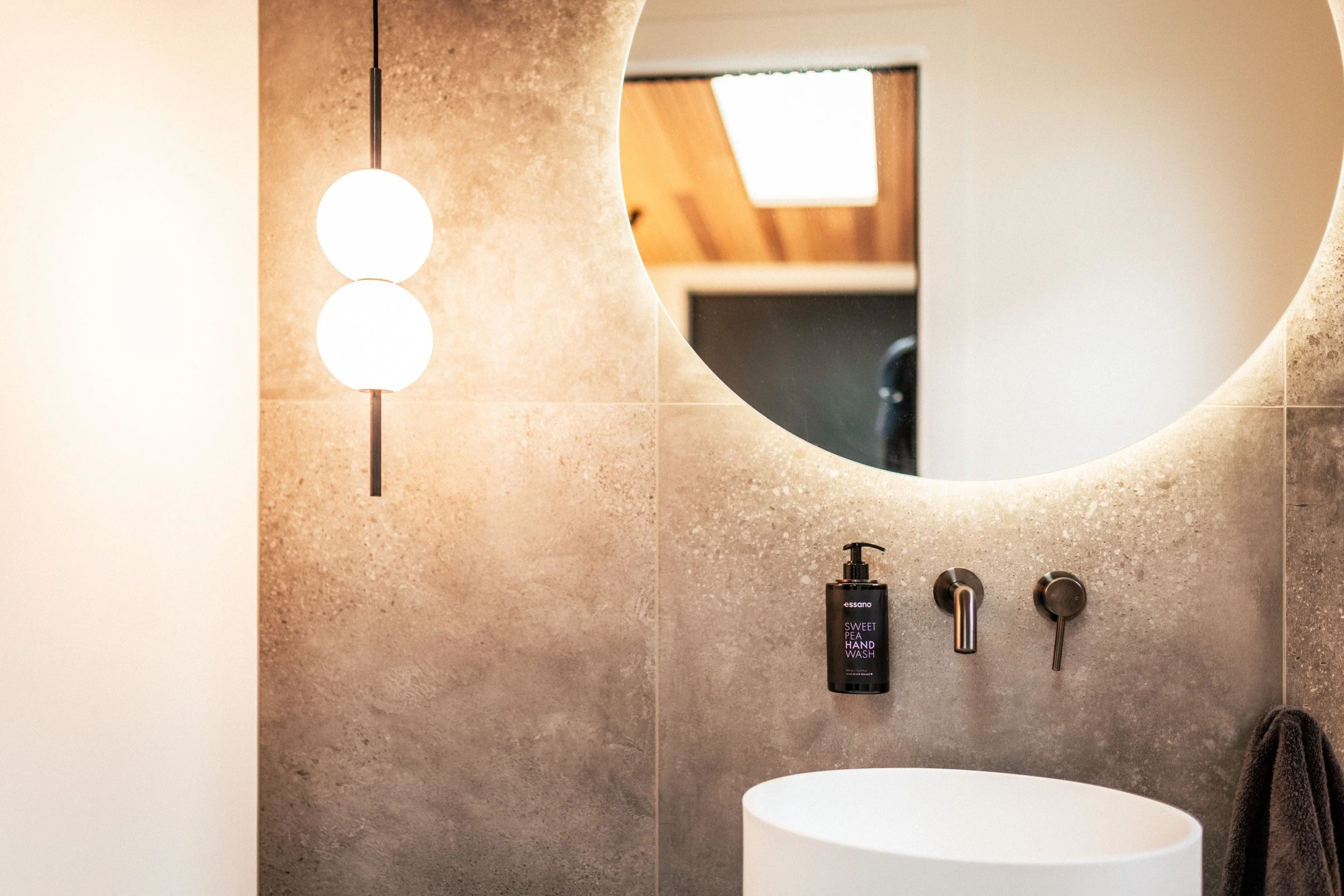 Modern powder room at Ocean View Te Arai featuring polished concrete flooring, concrete tiles, designer pendant lighting, and a large backlit mirror for a sleek, architectural finish.