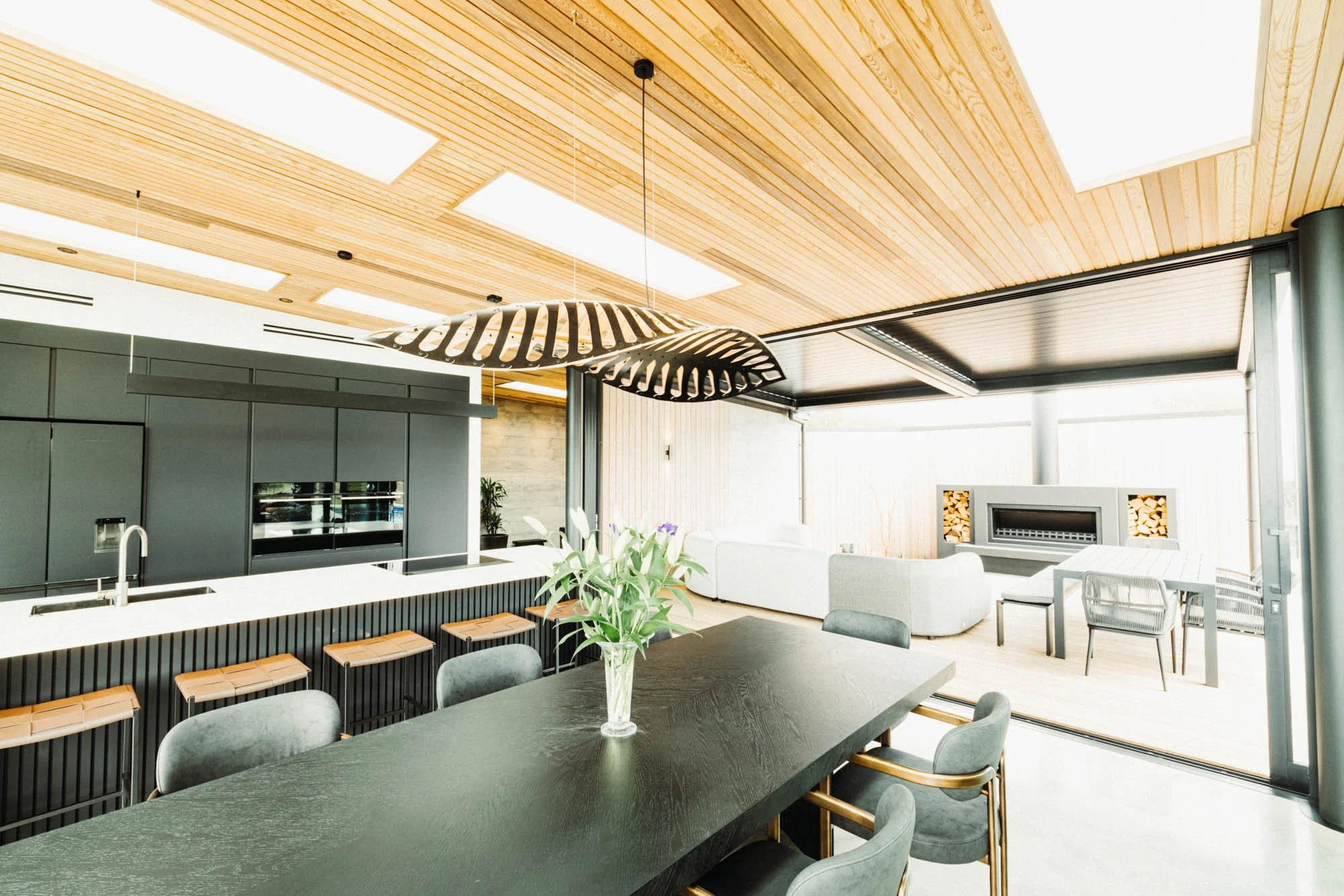 Interior view at Ocean View Te Arai showcasing the dining table with designer lighting, state-of-the-art kitchen island with bar stools, with floor-to-ceiling glass and sliding doors opening to the louvre-covered outdoor entertaining area and pool be