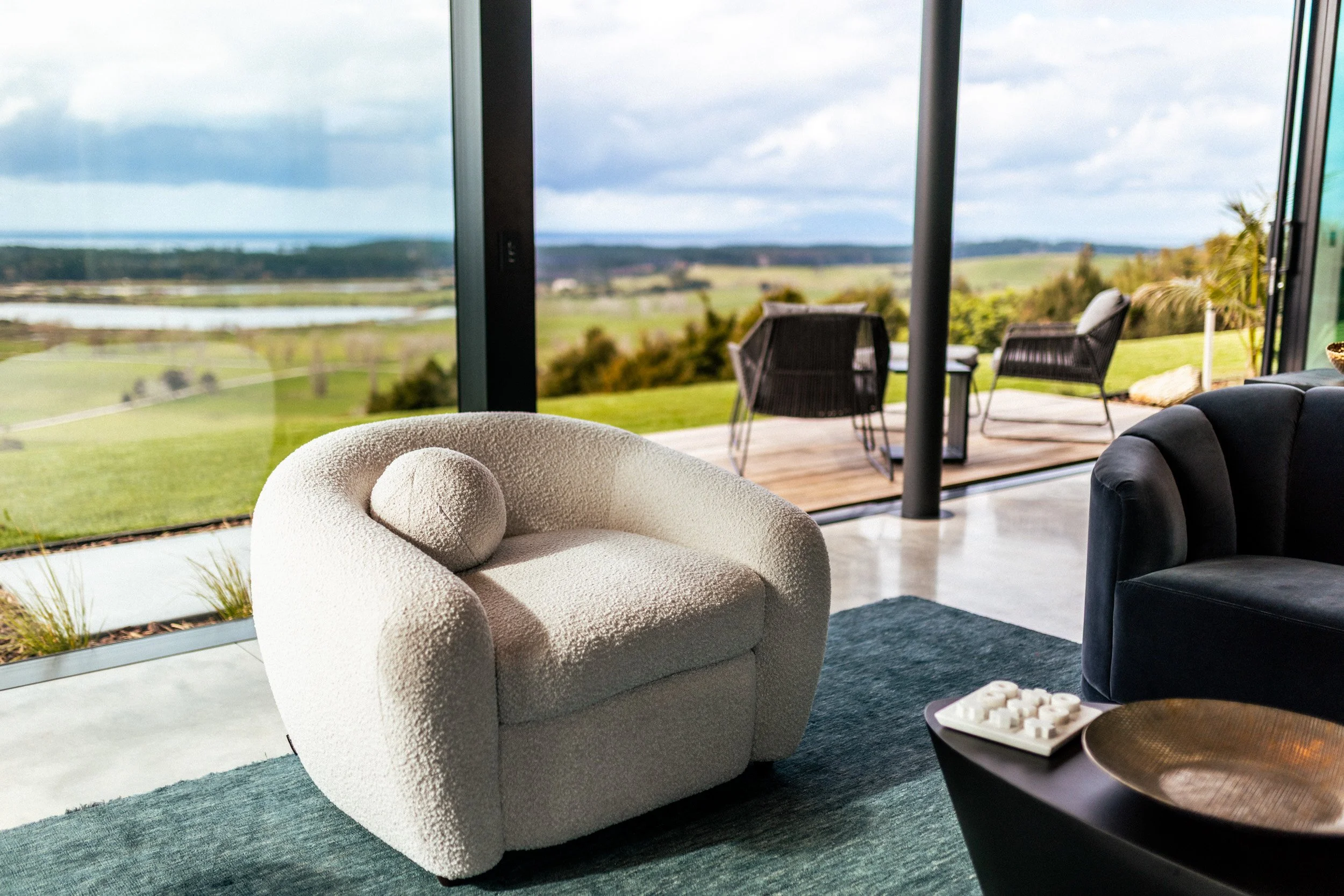 Designer occasional chair and luxurious rug in the living area at Ocean View Te Arai, with floor-to-ceiling glass framing expansive sea, lake, forest and coastal views.
