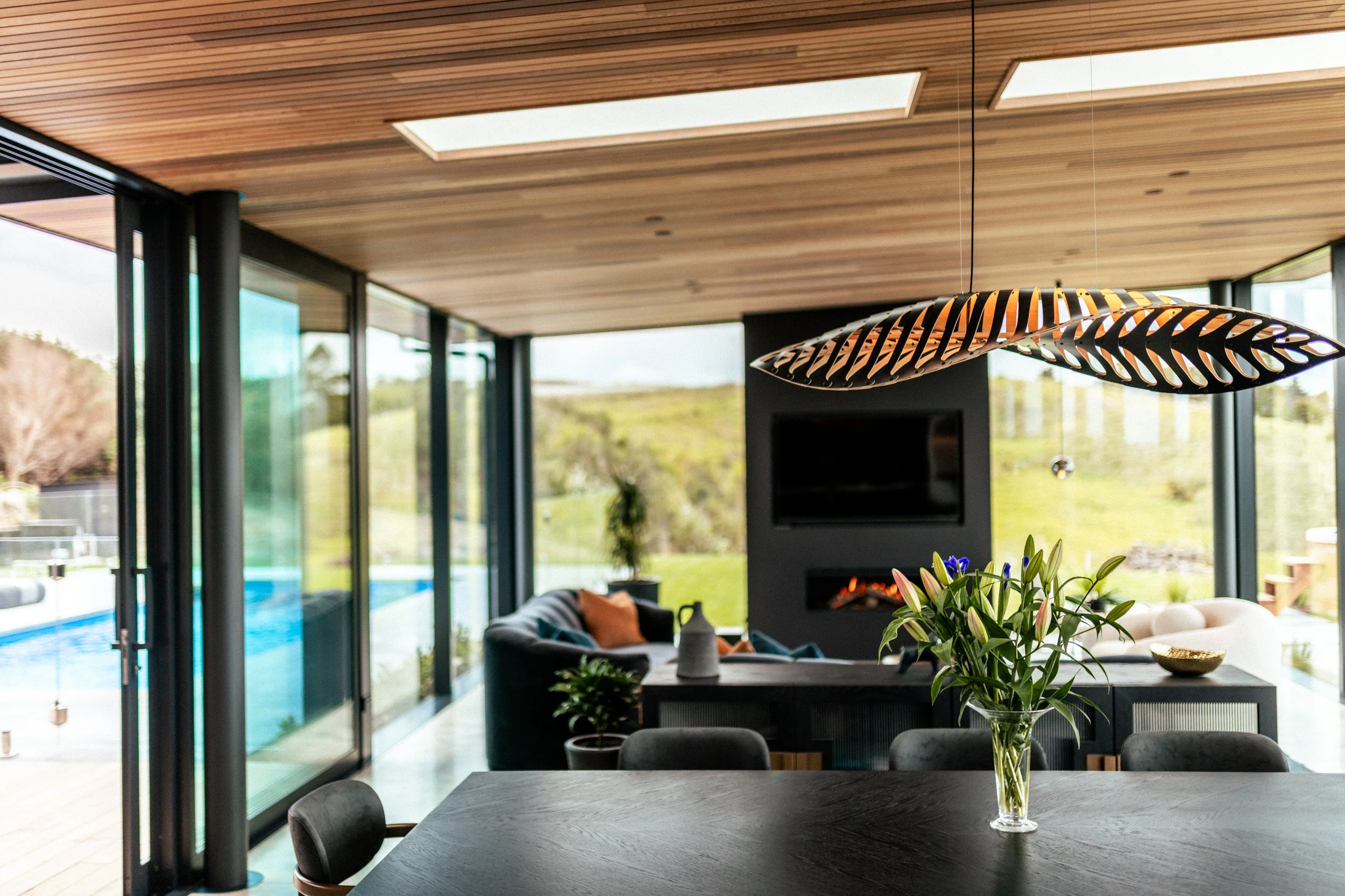 View over the designer dining table to the open-plan living area, with floor-to-ceiling glass on both sides framing the outdoor entertaining area, pool, spa, and expansive coastal views.