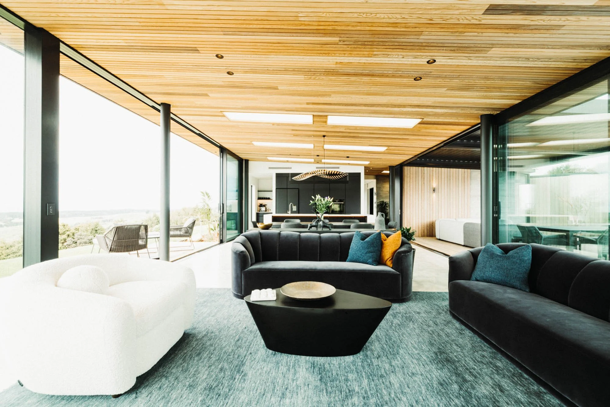 Open plan lounge, dining and kitchen area with floor-to-ceiling glass on both sides and designer furniture at Ocean View Te Arai, opening to sun-drenched decks and lawn that maximises breathtaking coastal views.