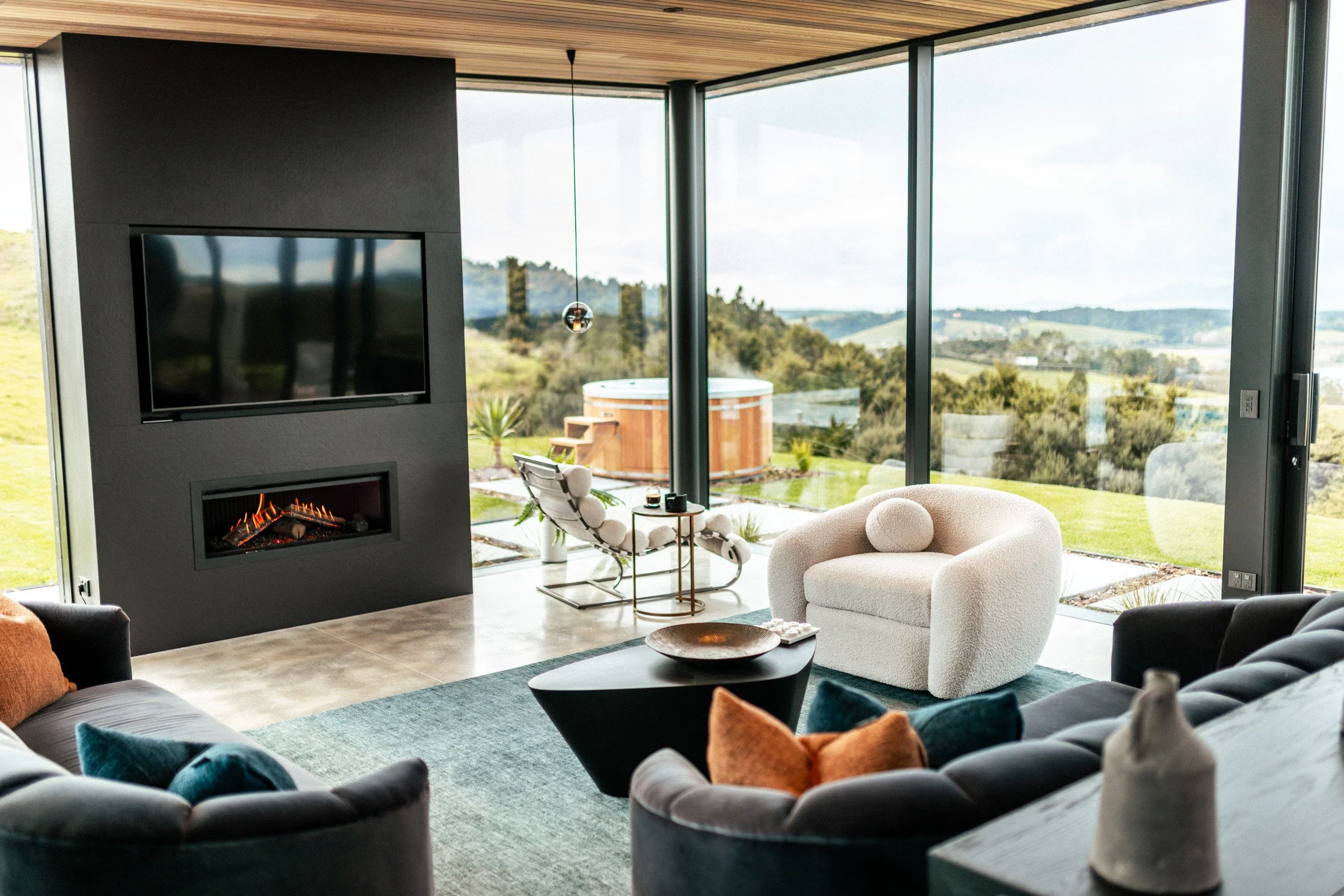 Lounge area with floor-to-ceiling glass, gas fireplace and designer furniture at Ocean View Te Arai, opening to sun-drenched decks, spa pool and lawn that maximises breathtaking coastal views.