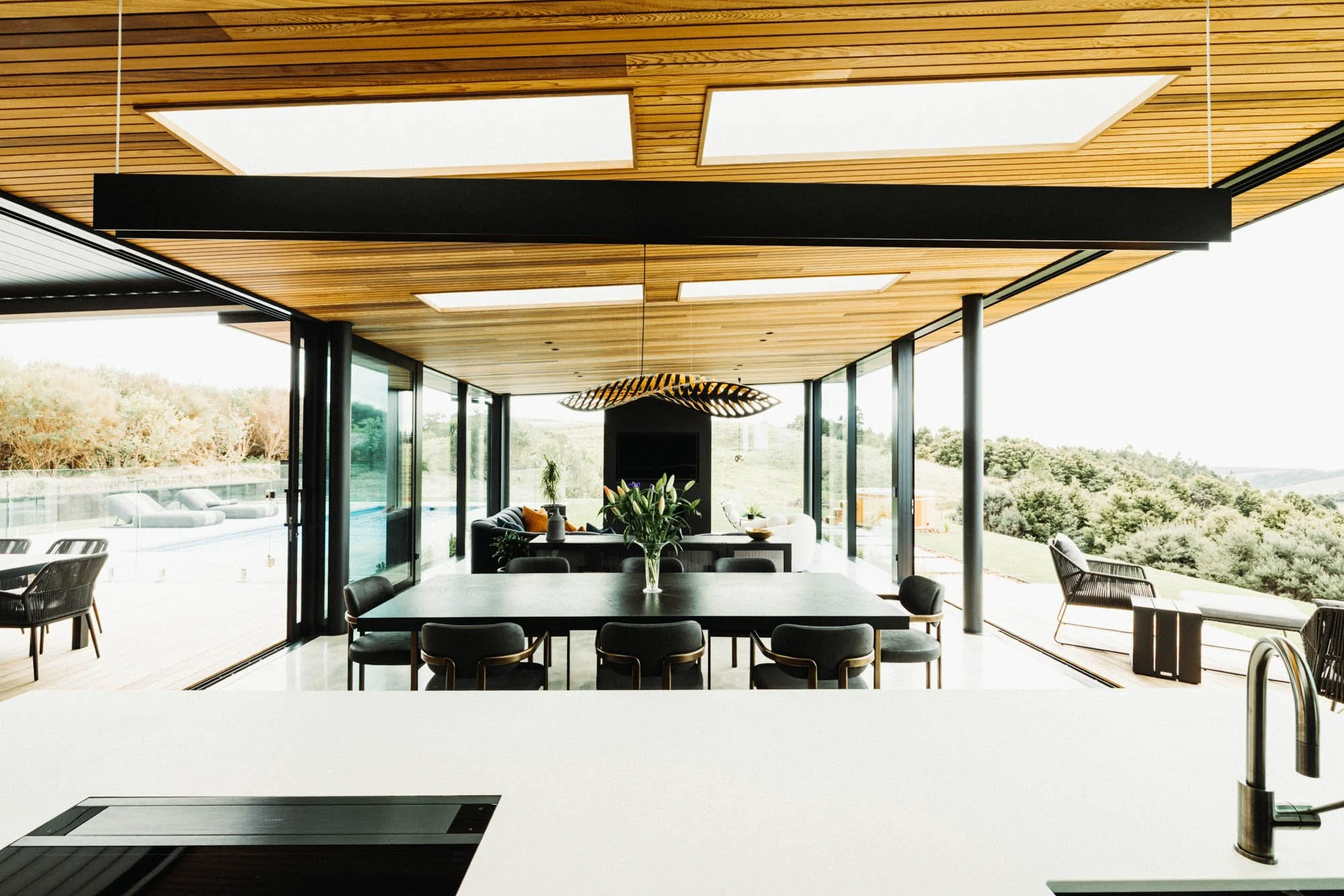 View over the concrete kitchen island at Ocean View Te Arai to the open-plan dining and living area, with floor-to-ceiling glass on both sides framing the outdoor entertaining area, pool, spa, and expansive coastal views.