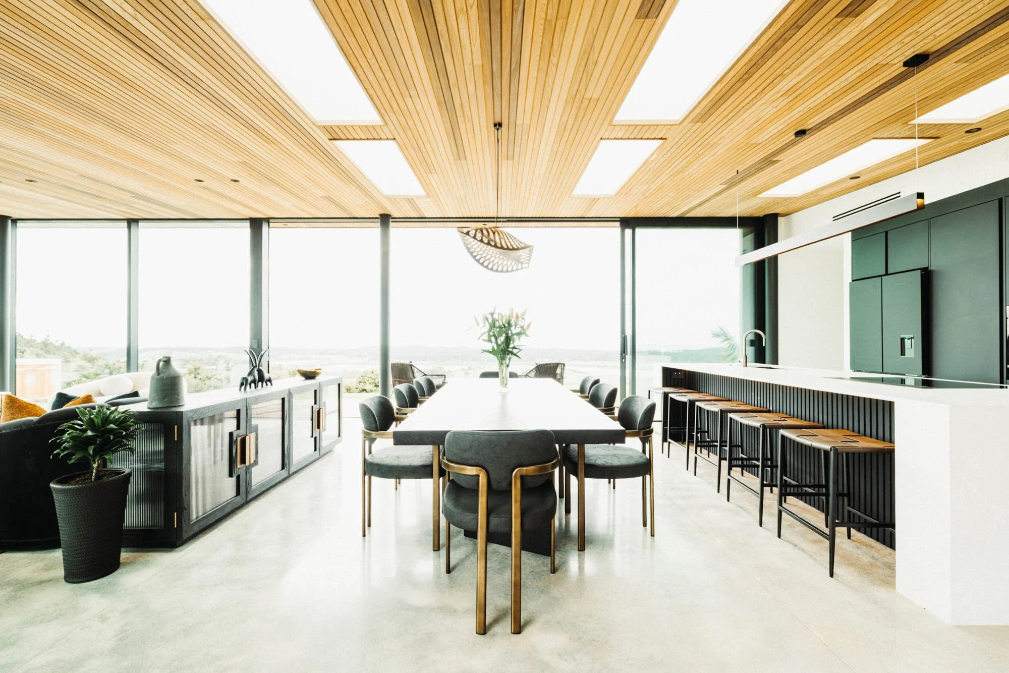 Interior view at Ocean View Te Arai showcasing the dining table with designer lighting, state-of-the-art kitchen island with bar stools, with floor-to-ceiling glass and sliding doors opening to the expansive ocean, lake, forest and farmland views.