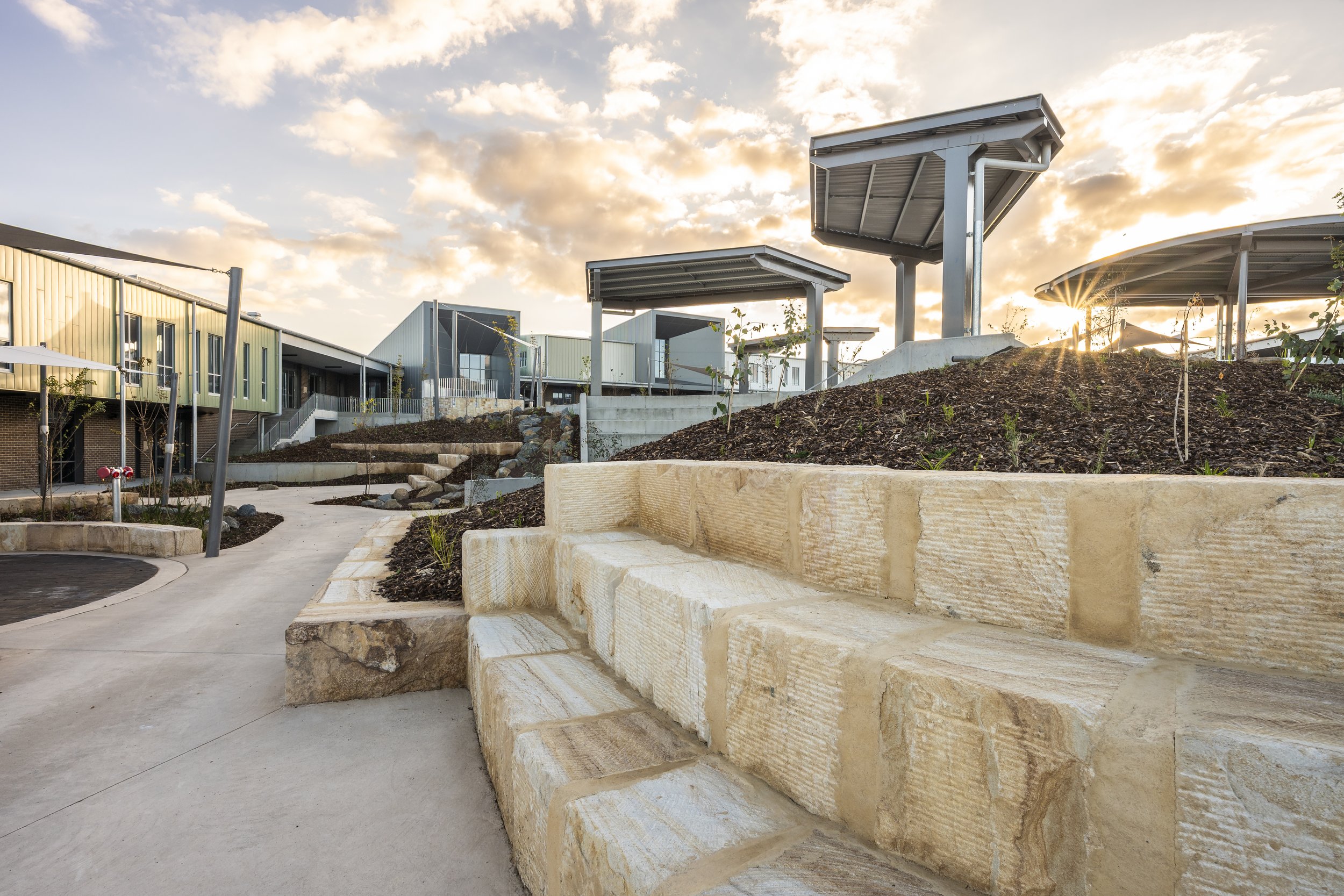 Modern building with green and gray exterior and a landscaped outdoor area with concrete stairs and wooden seating, under a sky with scattered clouds at sunset.