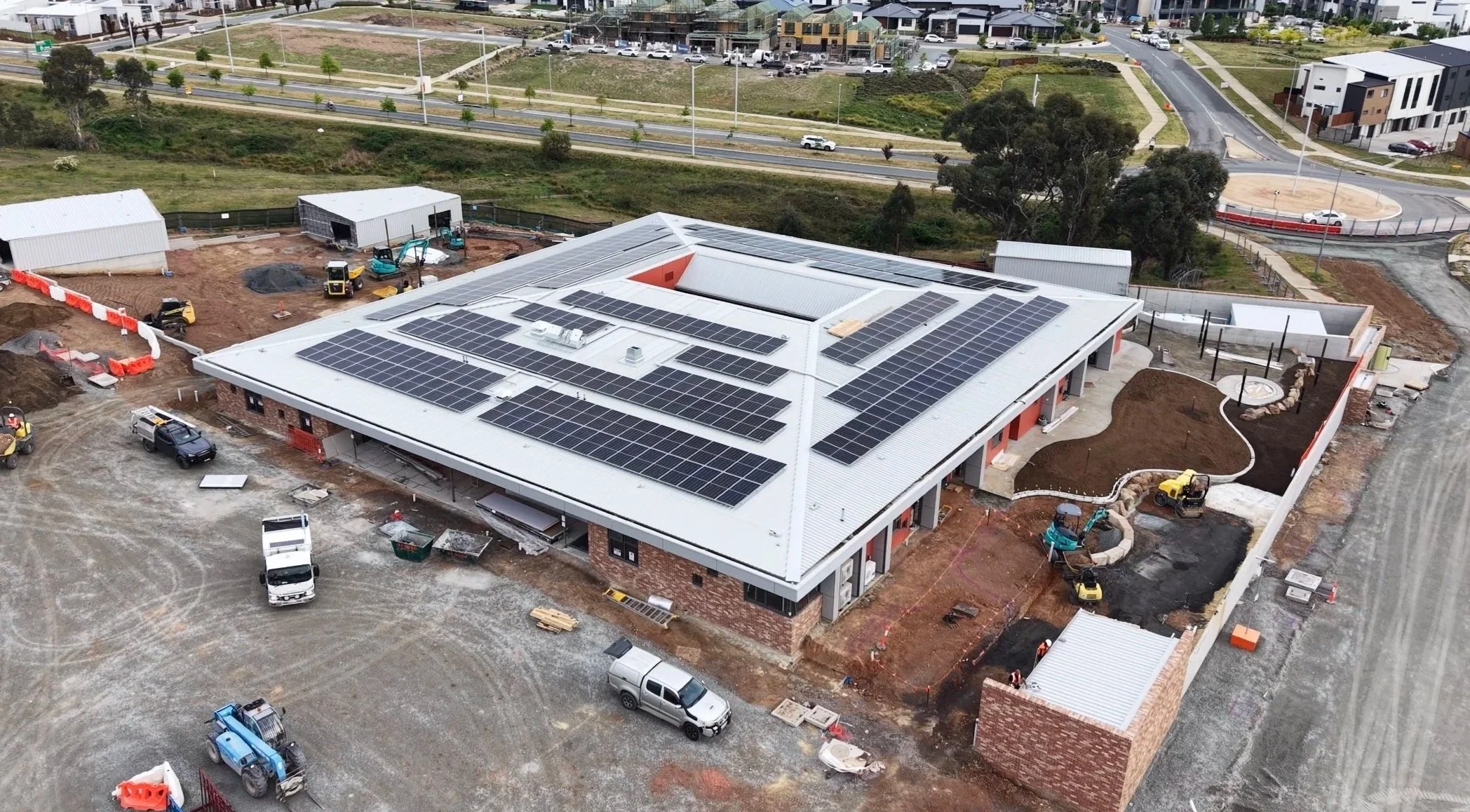 An aerial view of a construction site with a building featuring solar panels on the roof, surrounded by construction equipment, vehicles, and landscaping in progress.
