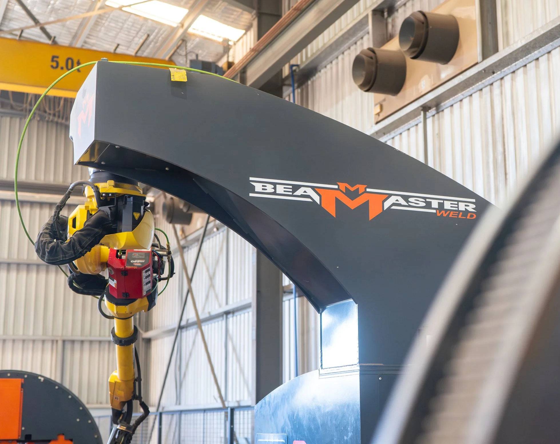 A robotic welding arm is welding a large metal structure inside an industrial warehouse. The arm is yellow with black gloves on the operator's hand, and the structure has the logo 'BEAMASTER WELD' on it.