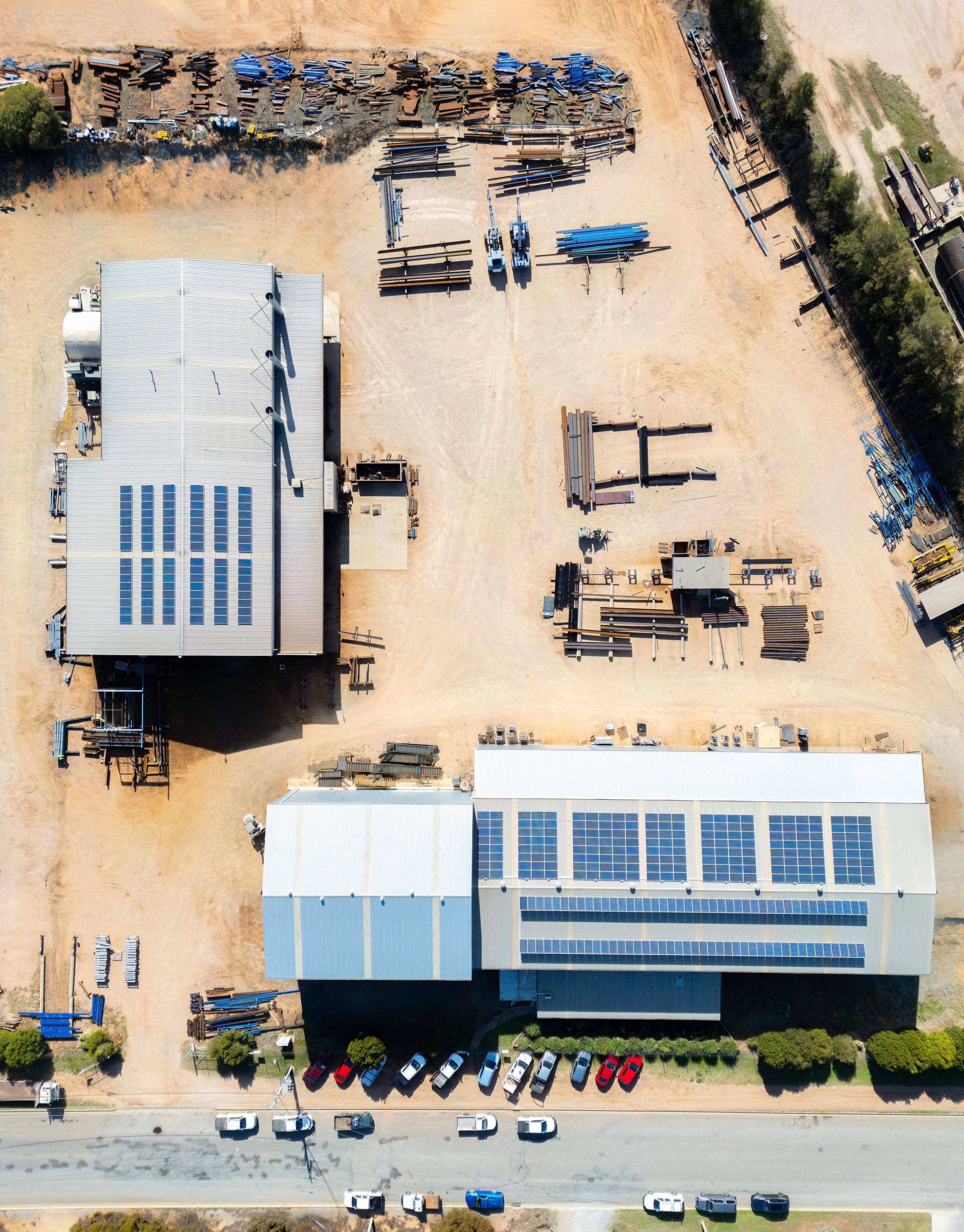 An aerial view of an industrial area with large warehouse buildings, parking lot, and stacks of materials and equipment.