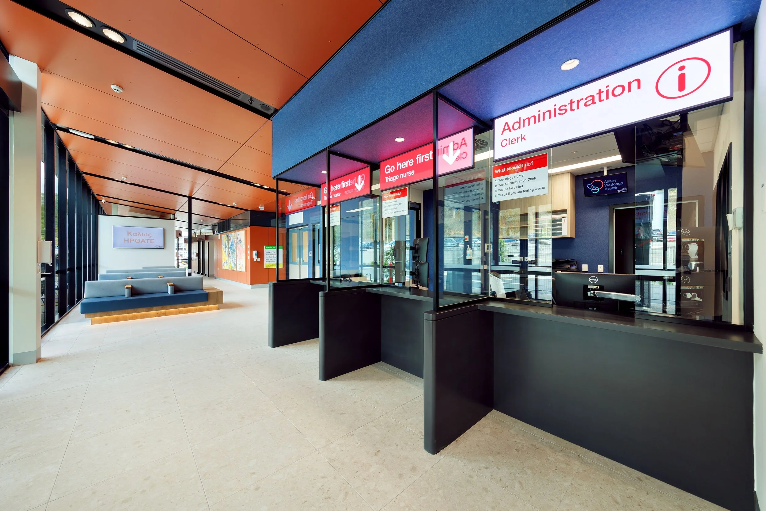 Hospital reception area with a black reception desk, digital screens, and orange and blue ceiling panels. Signs indicate the administration desk and triage nurse station. There are seating areas with gray benches and a door with the Albury Wodonga Health logo.
