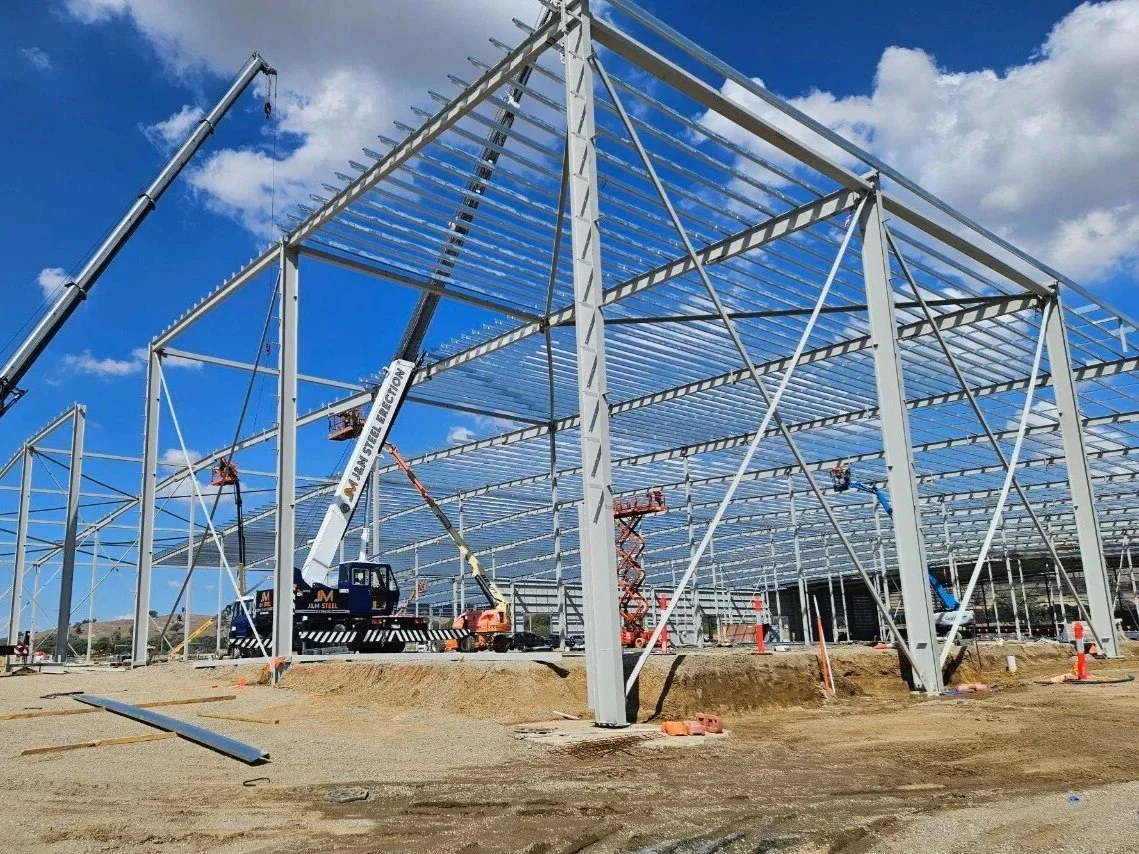 Construction site with a large metal framework under blue sky with clouds, cranes, construction equipment, and partially built structure.