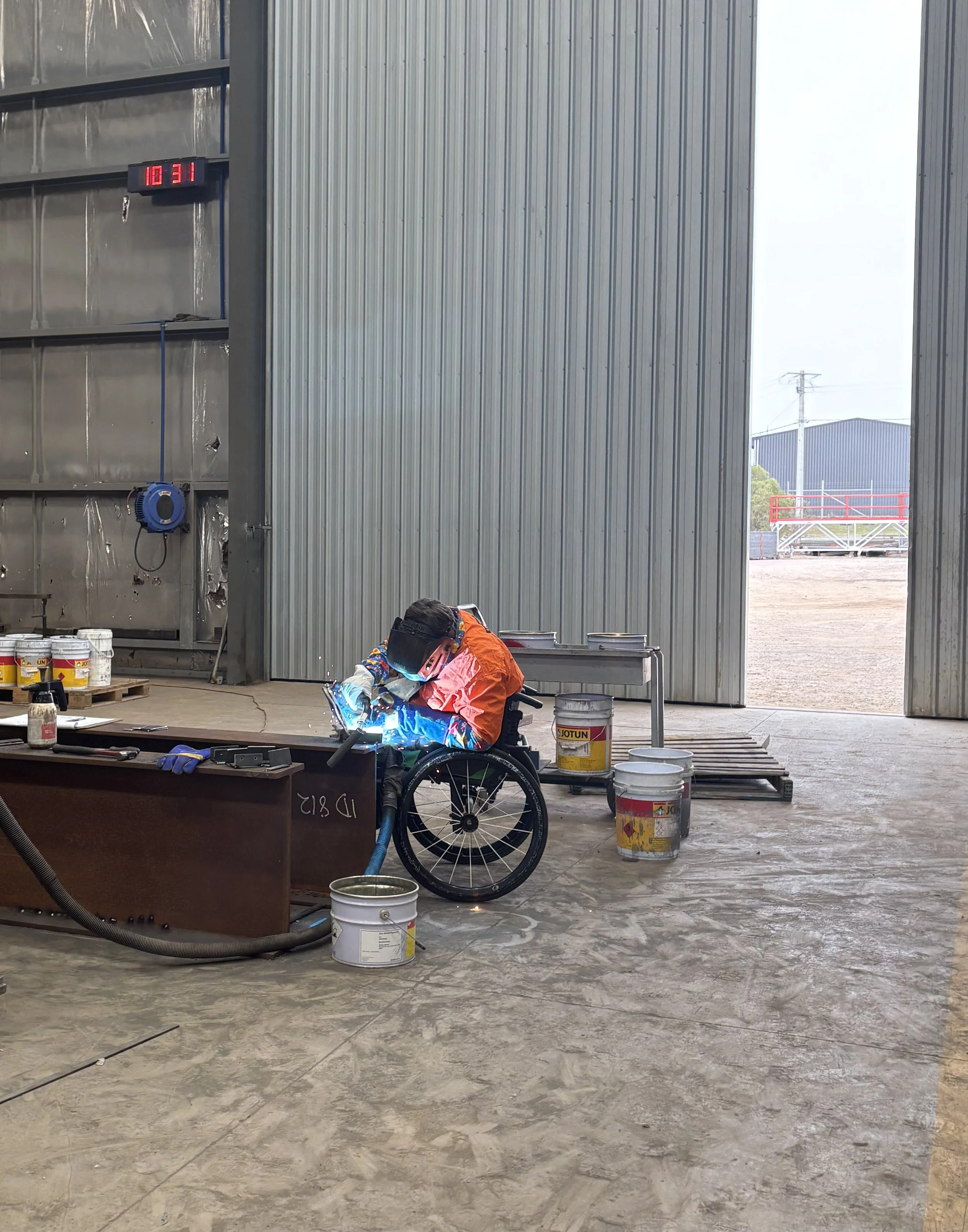 In the workshop, a work experience student in a wheelchair is welding together some plates.