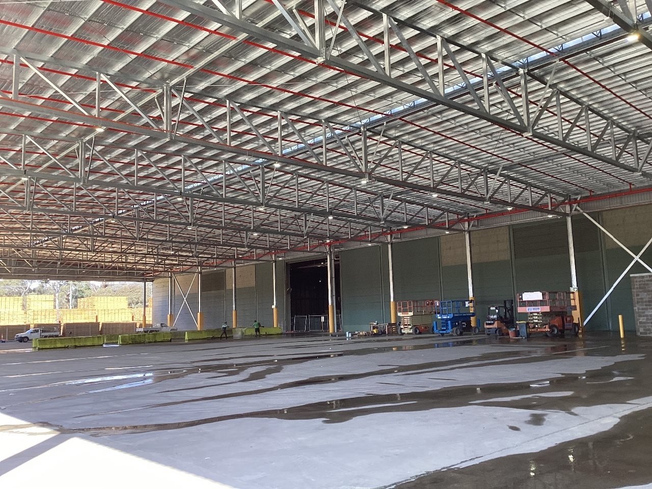 Inside of a large warehouse or hangar under construction, with visible metal roof trusses, a partially wet concrete floor, and construction equipment and workers in the background.