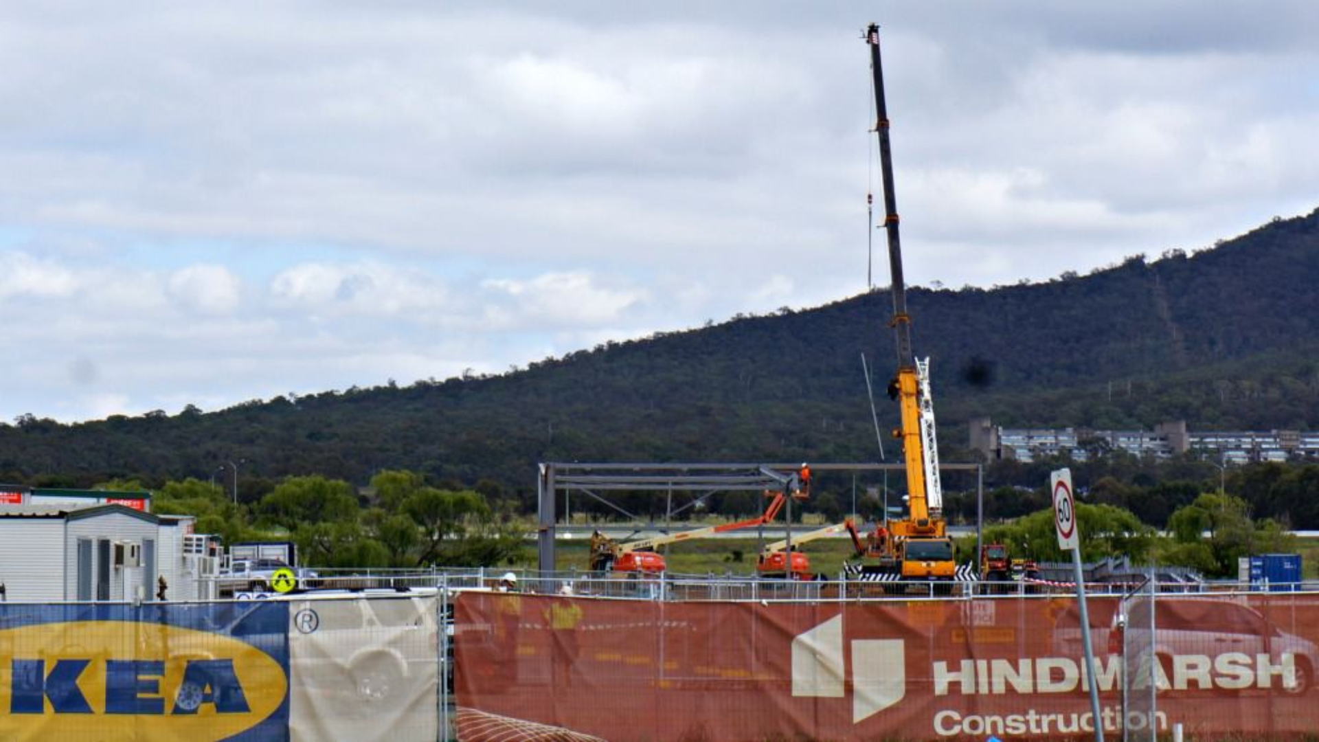 Construction site with a large mobile crane lifting a steel framework, with a mountain and cloudy sky in the background, and advertising banners in the foreground.