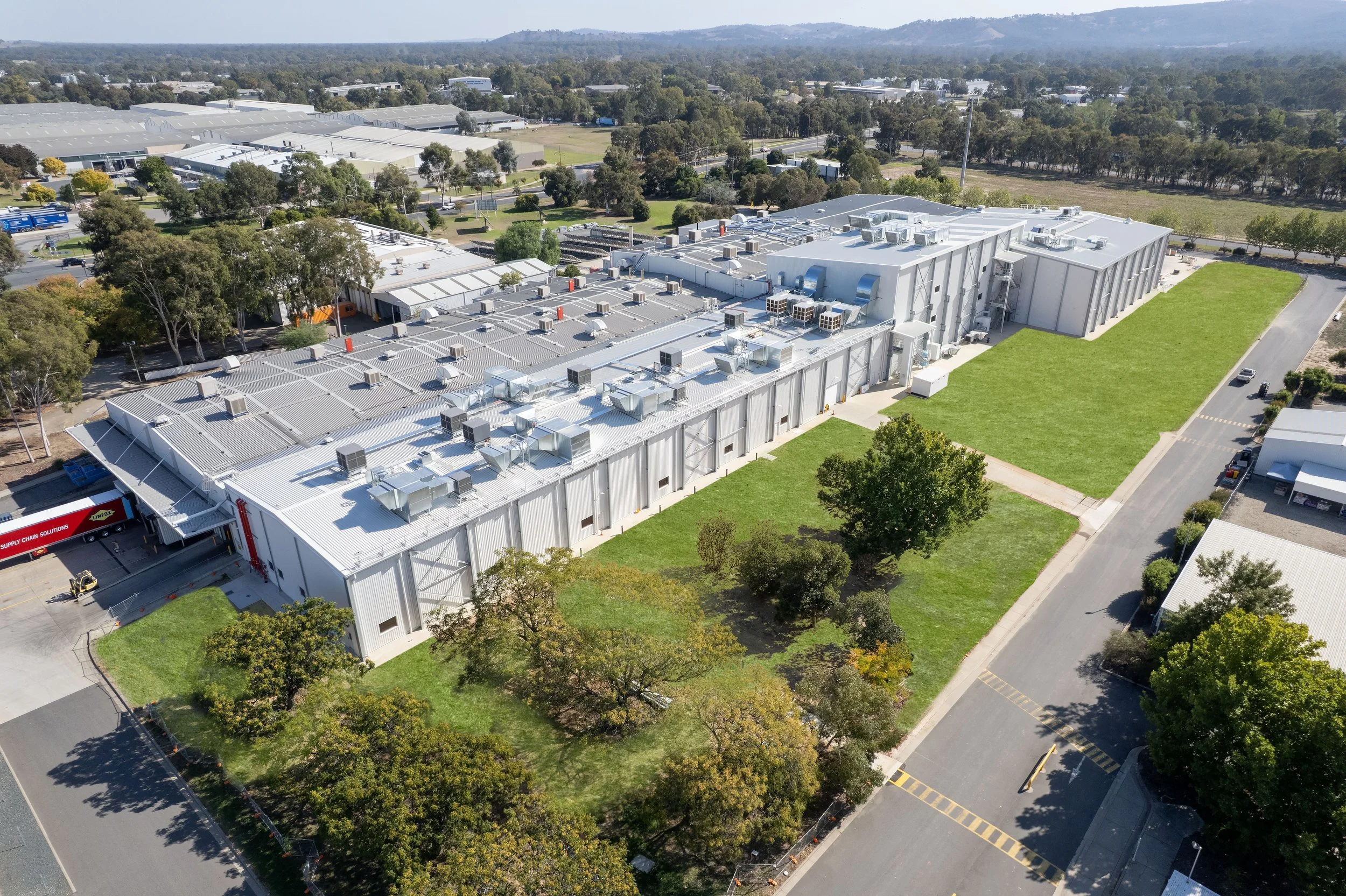 Aerial view of a large industrial building with a flat metal roof equipped with numerous HVAC units and solar panels, surrounded by green lawns, trees, and roads, with other industrial buildings and trees in the background.