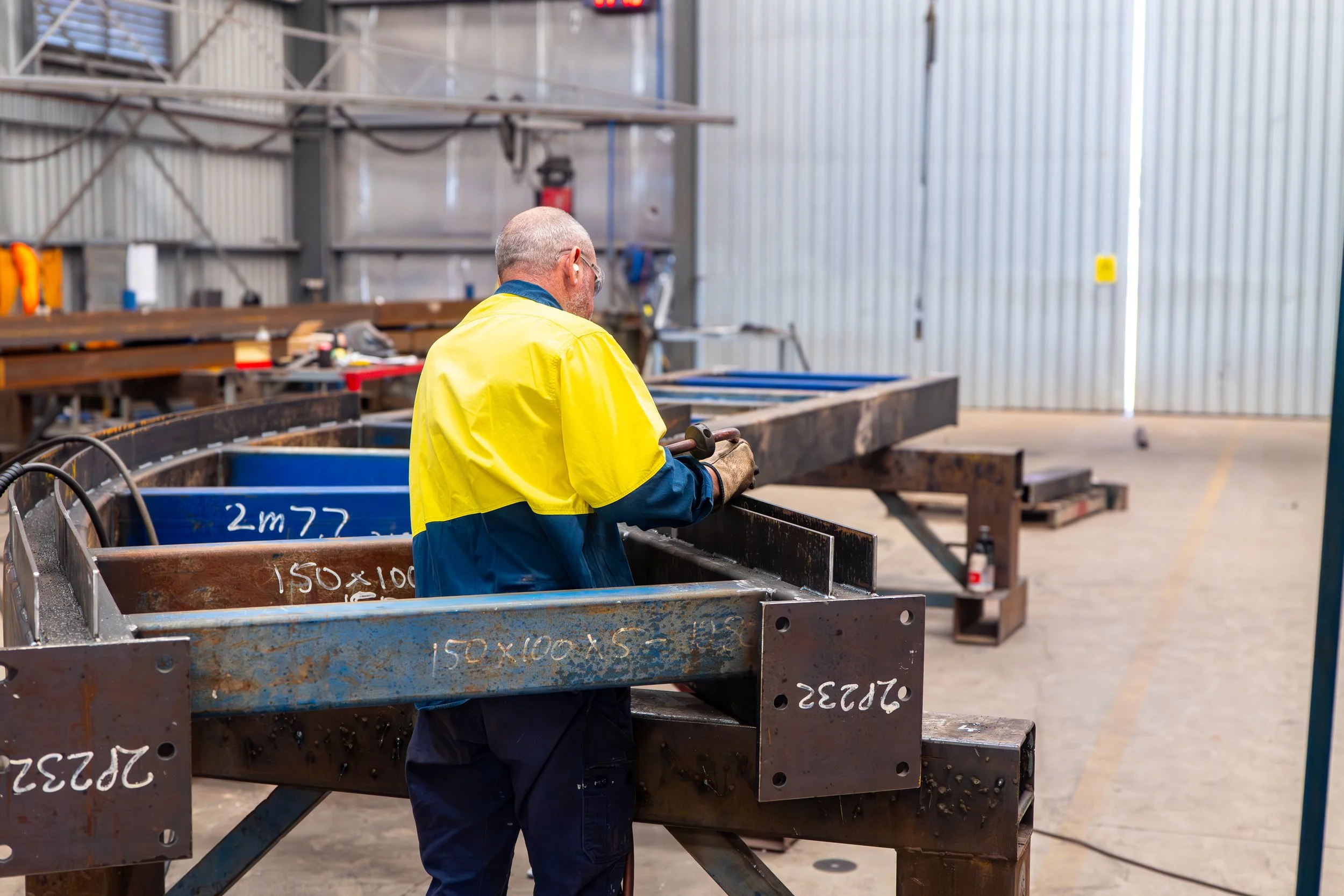 A worker wearing safety glasses and a yellow and blue safety jacket inspects metal beams inside a large industrial warehouse.
