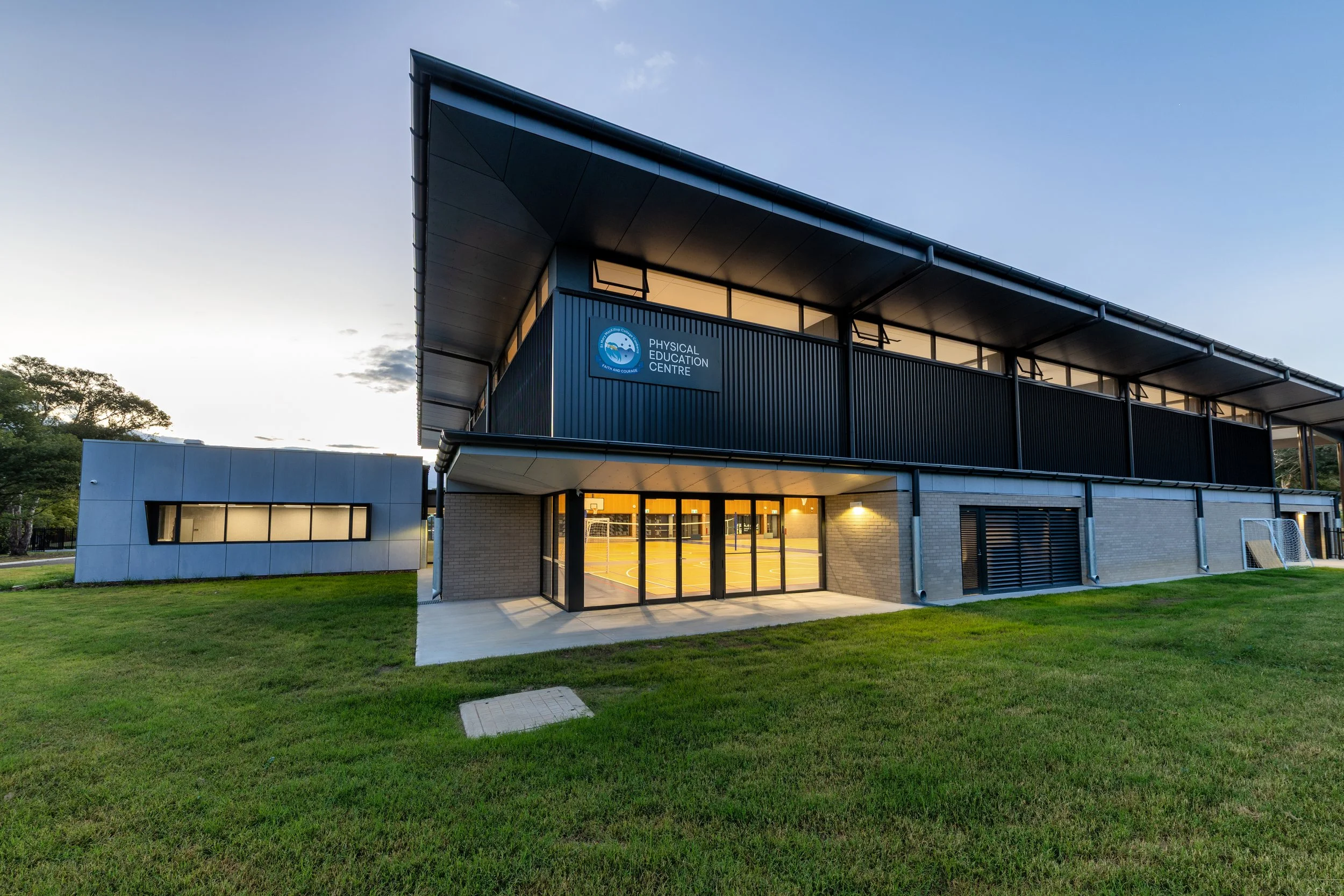 Modern building with a blue sign reading "Physical Education Centre" and an outdoor sports court visible through glass doors, surrounded by a well-kept grassy area.