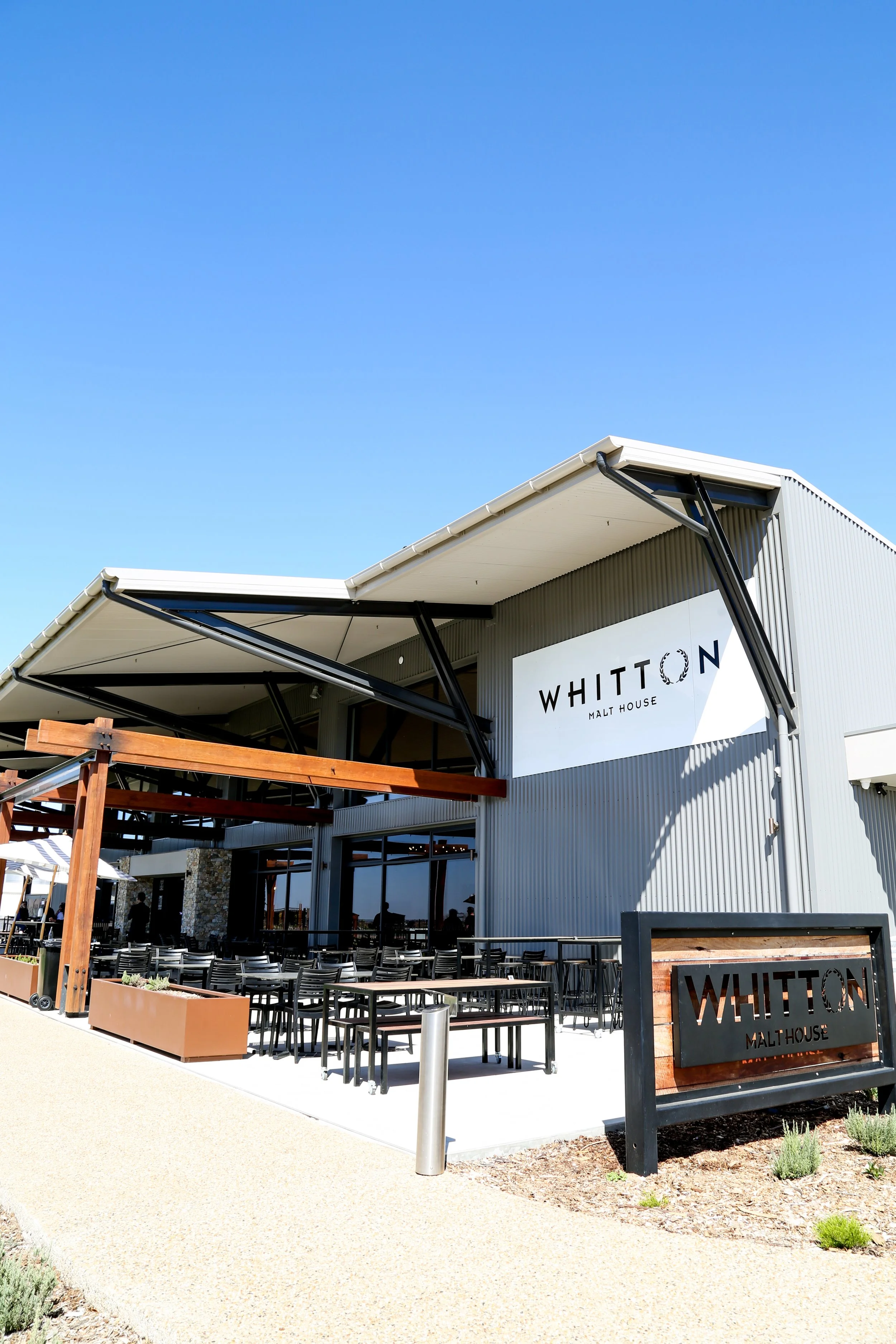 Exterior of WiTTON Malt House restaurant with outdoor seating, black tables and chairs, a wooden pergola, and clear blue sky.