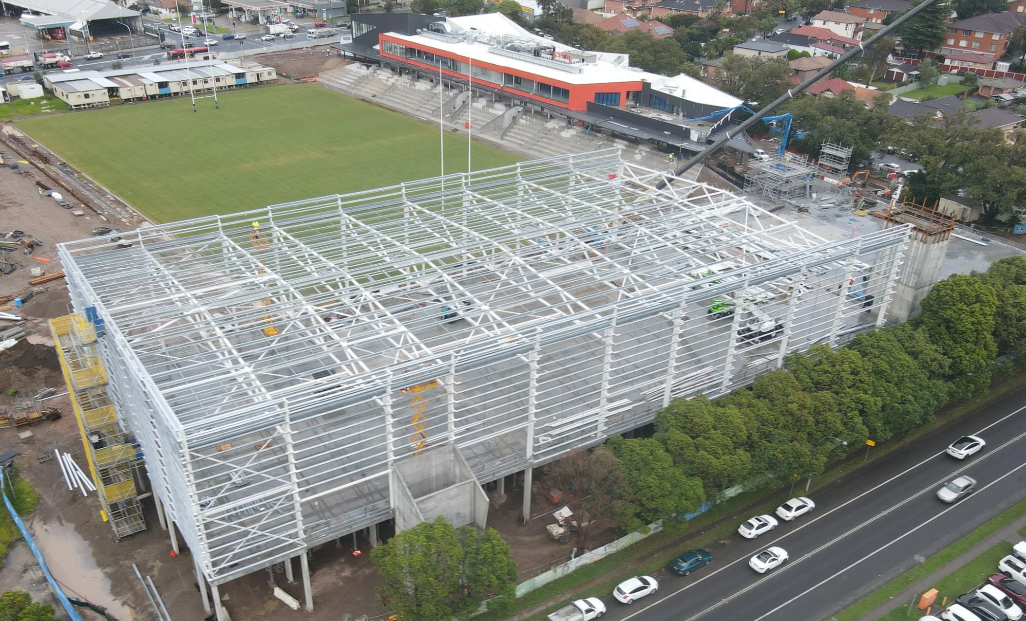 Aerial view of a construction site where a large modern building with a metal frame is being constructed, adjacent to a football field and nearby residential area.