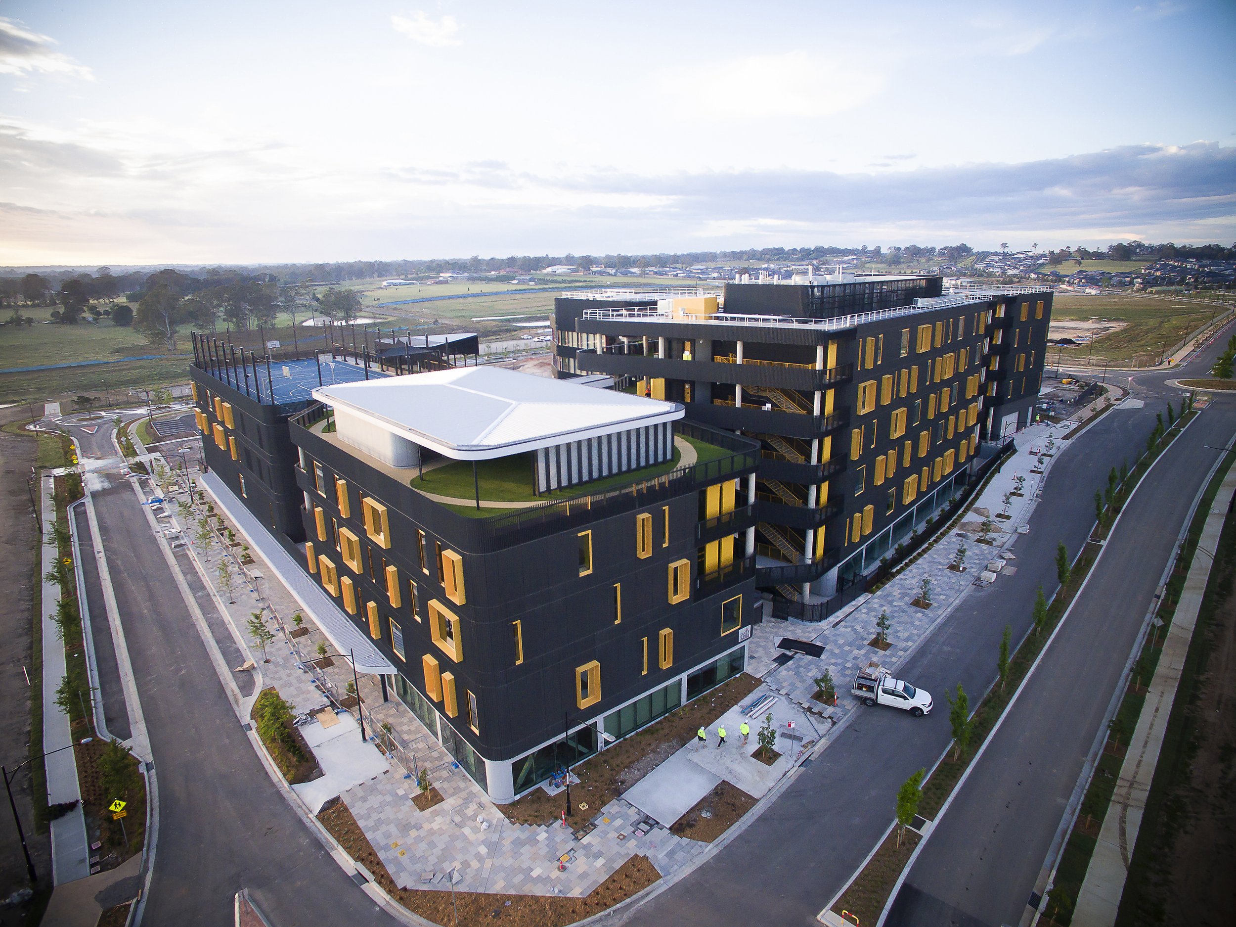 A multi-story modern building with black and yellow exterior, rooftop sports court, and surrounding streets and sidewalks.