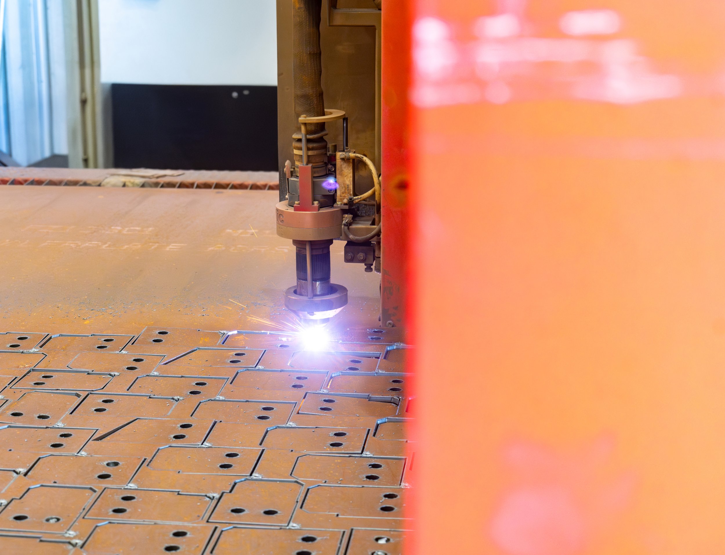 A close-up of a plasma cutter cutting through metal, with sparks and bright light visible at the point of contact. The background includes industrial elements and part of a workspace.