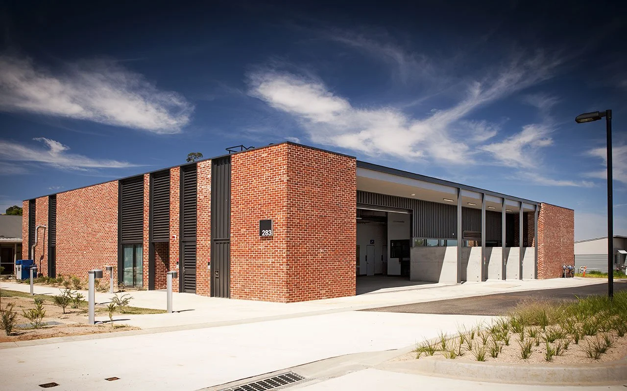 Modern building with red brick exterior, metal accents, and a parking area under a partly cloudy sky.