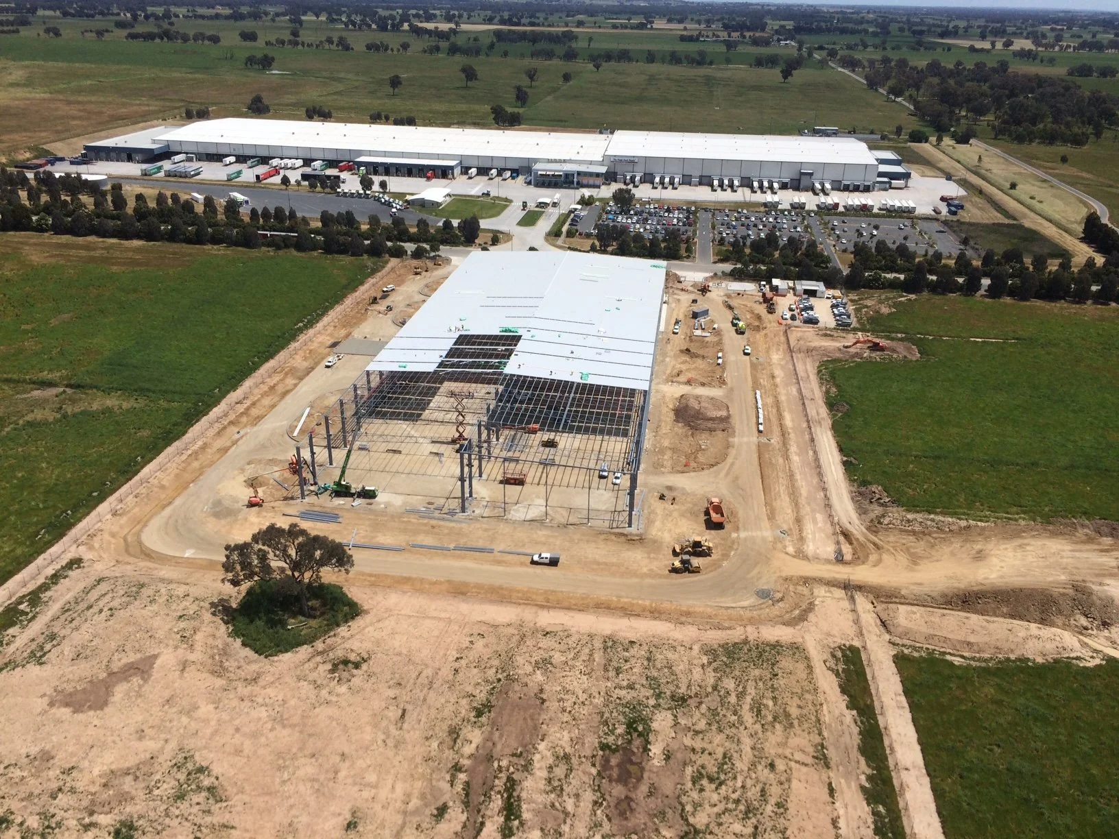 Aerial view of a construction site for a large warehouse or distribution center, with the steel framework in place and roofing being installed, surrounded by open land and a parking lot.