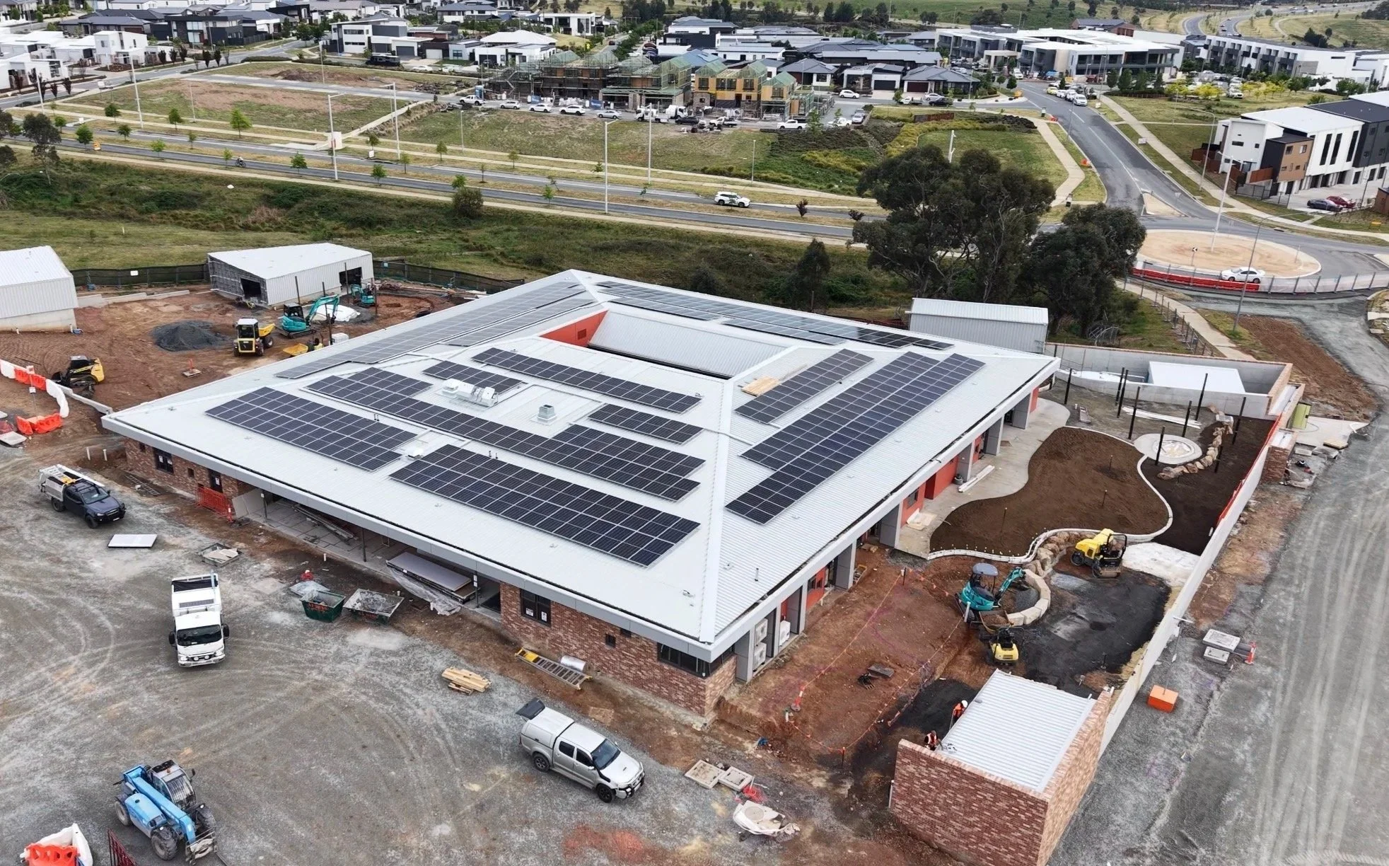 Construction site with a large building featuring solar panels on the roof, surrounded by ongoing ground work, construction vehicles, and nearby residential houses and roads.
