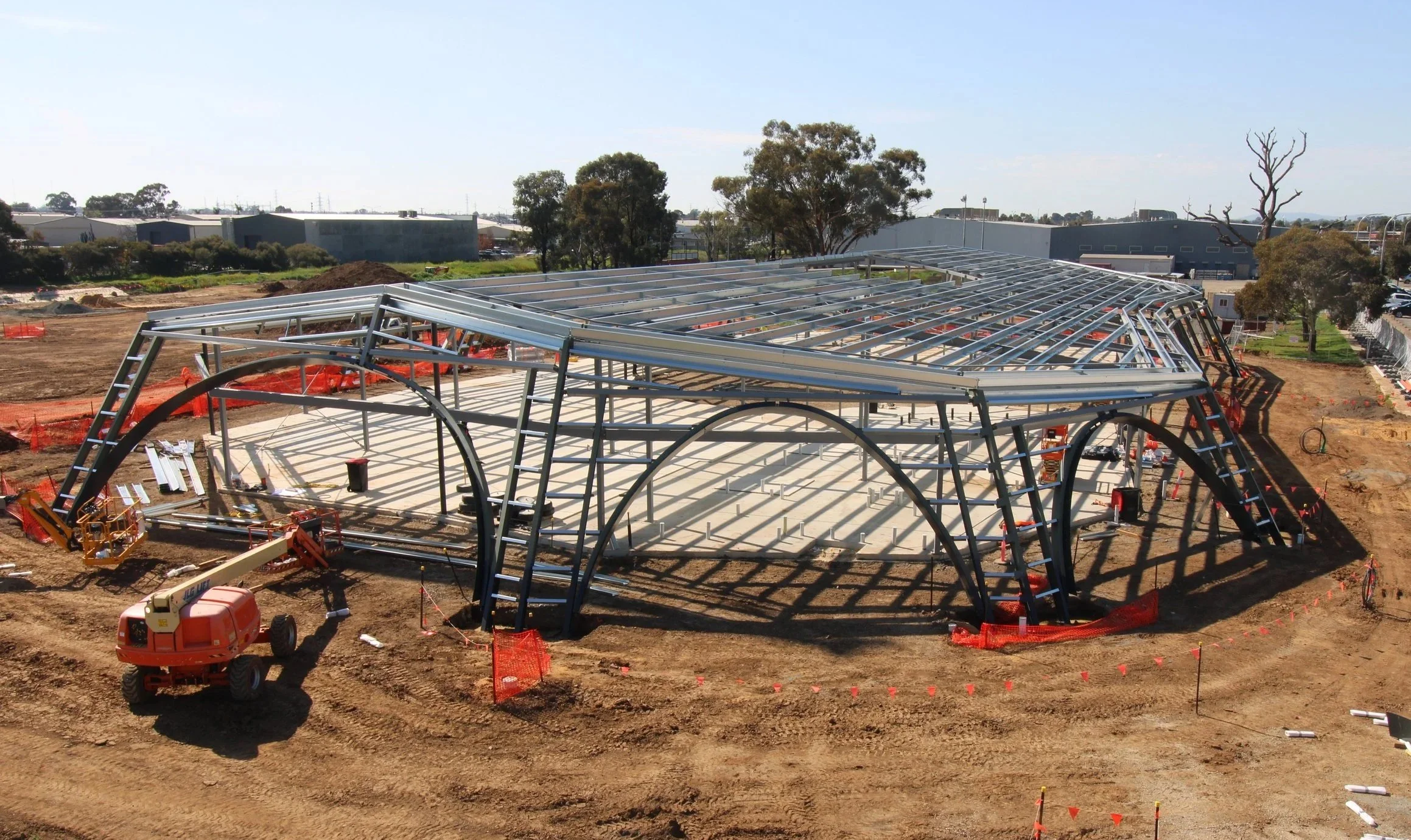 Under construction site featuring a metal framework structure, construction equipment, and dirt ground with orange safety barriers and fencing.