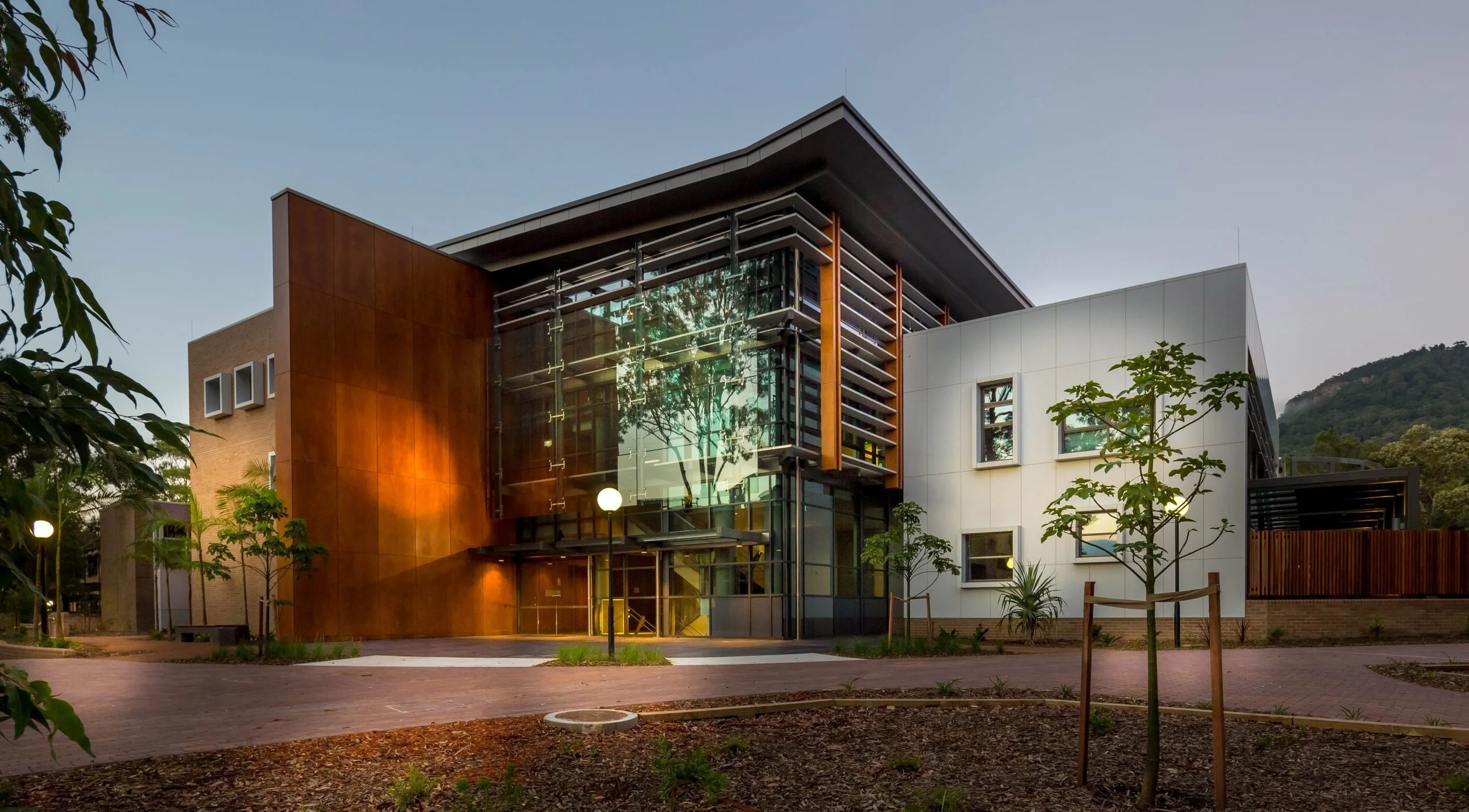 Modern multi-story building with glass walls, wooden panels, and white exterior walls, surrounded by small trees and lampposts, with a hill in the background at dusk.