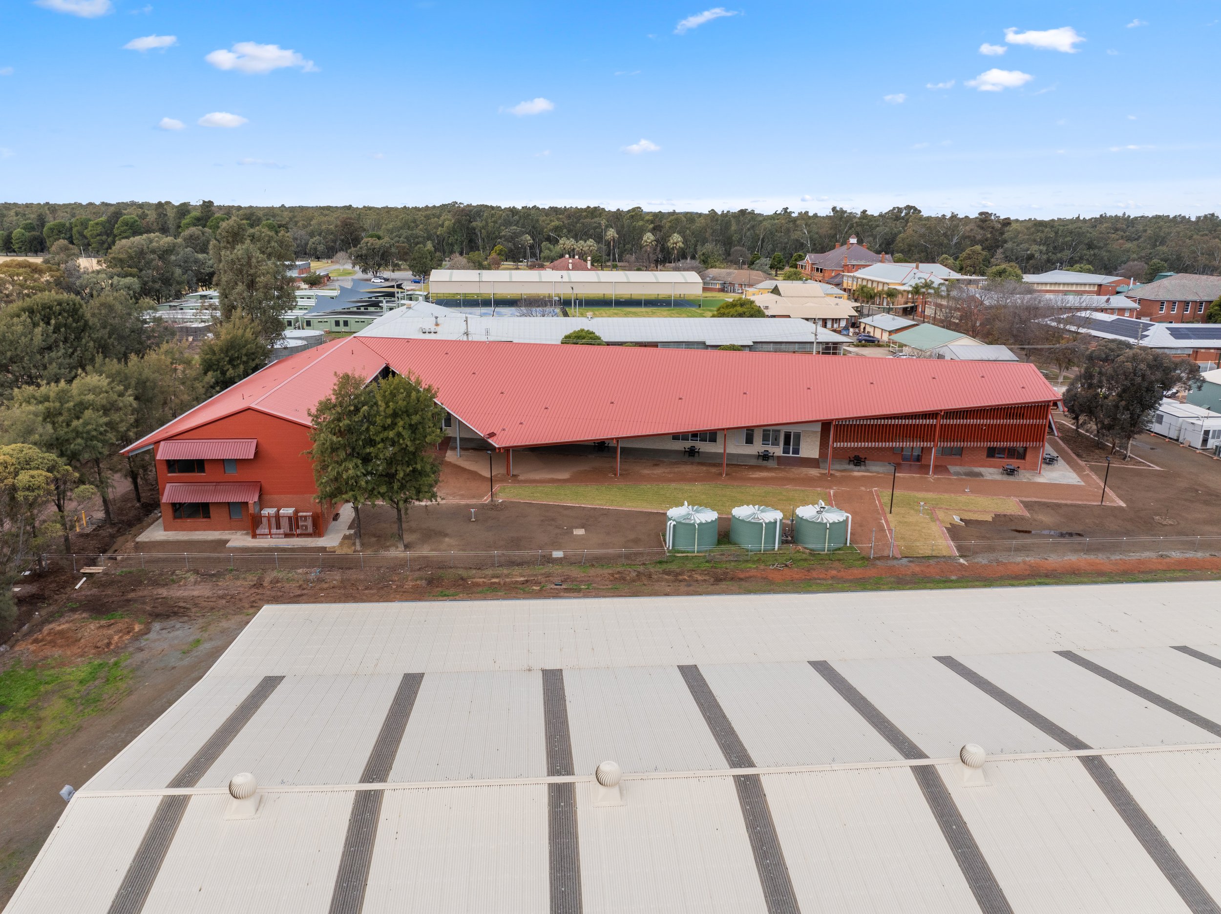 Aerial view of a campus with a large white-roofed building in the foreground, some trees, and multiple smaller buildings with various roof colors beyond. The background includes a forest and a blue sky with scattered clouds.