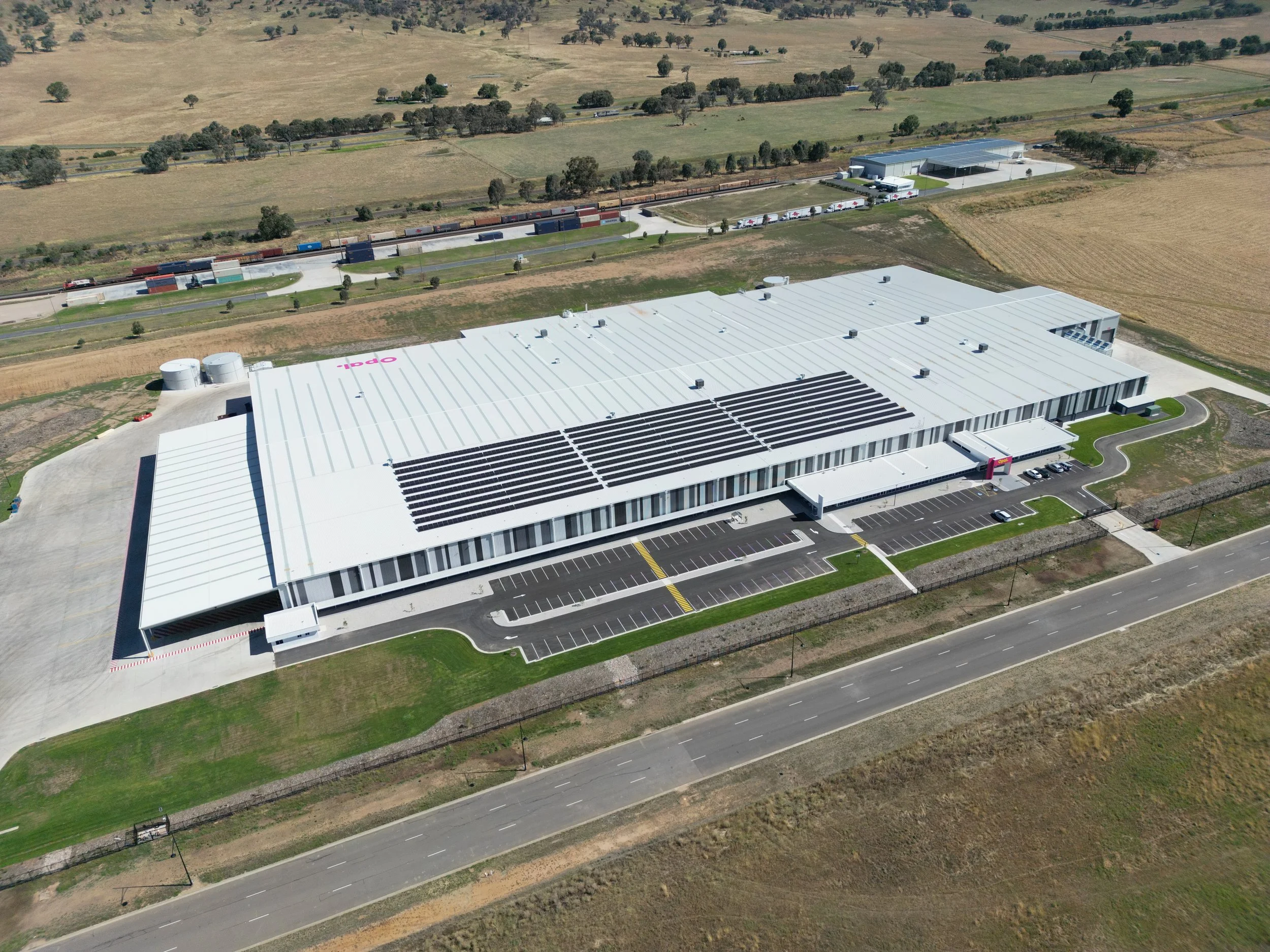 An aerial view of a large industrial building with solar panels on the roof, a parking lot, and a rural landscape with fields and trees in the background. A freight train runs parallel to the building.
