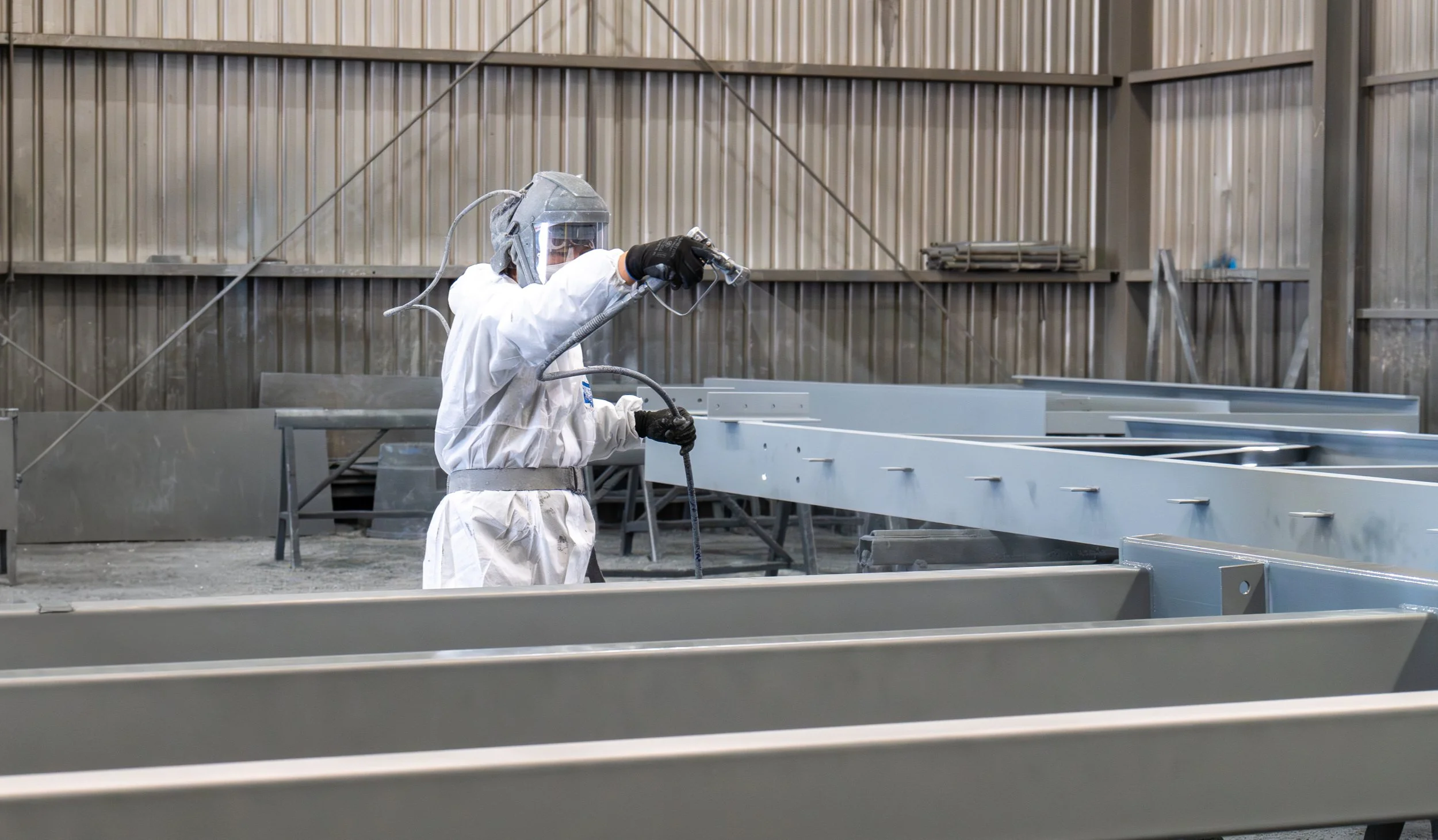 A worker in protective gear using a spray gun on metal framework inside an industrial facility.