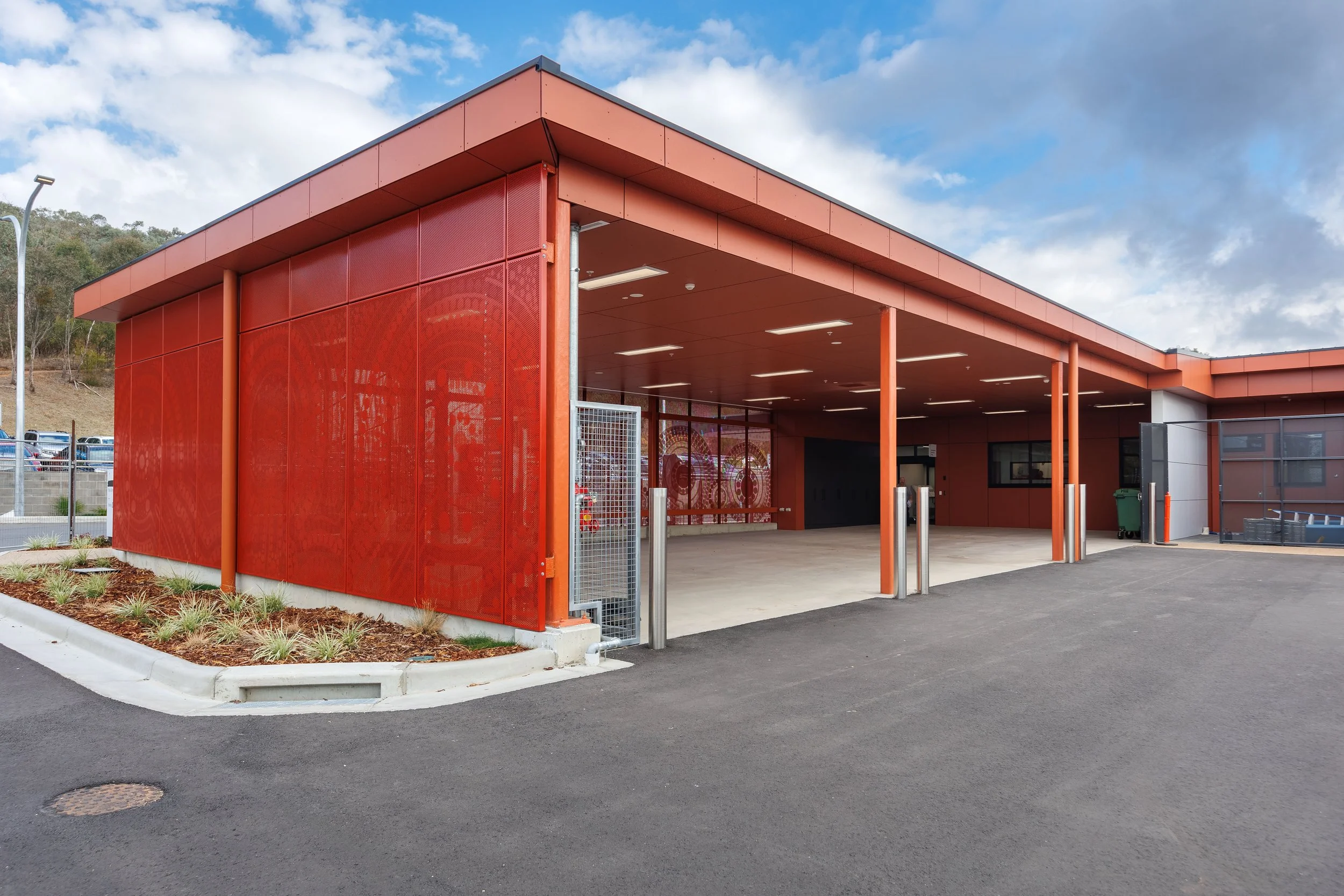 Modern orange building with open carport, surrounded by parking lot and landscaping, under partly cloudy sky.